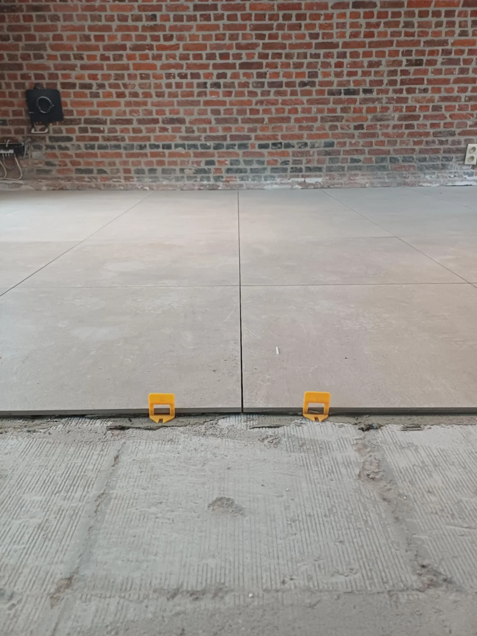 Tegels worden gelegd op de vloer voor een vloerinstallatie, met behulp van gele klemmen voor de positionering, tegenover een bakstenen muur.