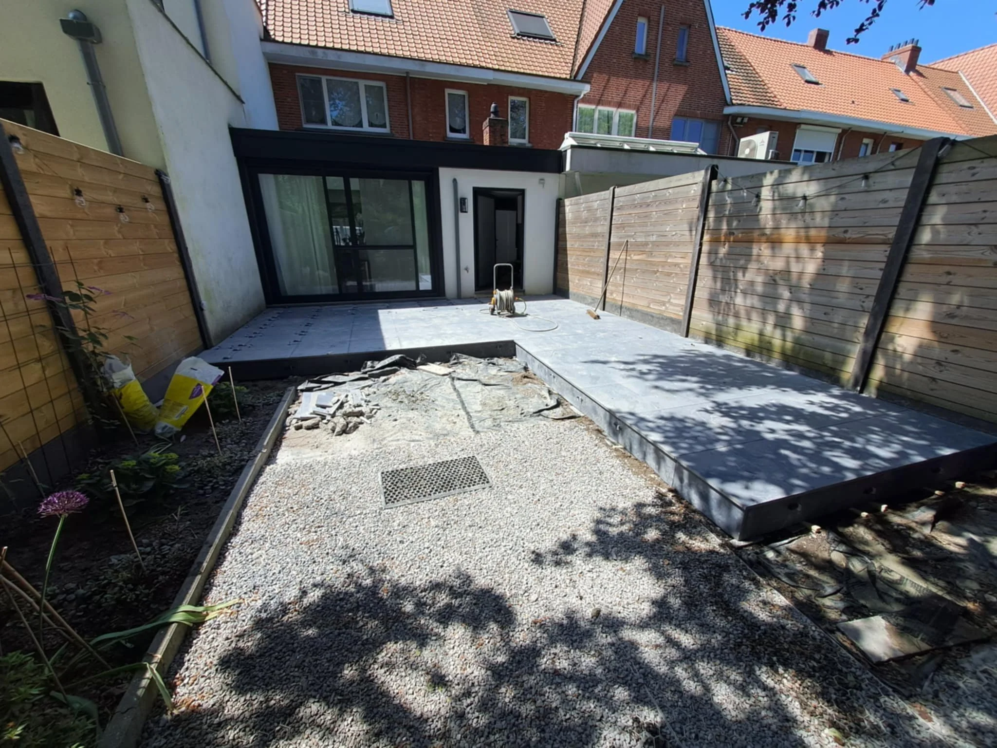 Tuin met een betegeld terras en een rondom houten schutting, achter een modern huis met grote ramen, met gereedschap en bouwmaterialen aanwezig.