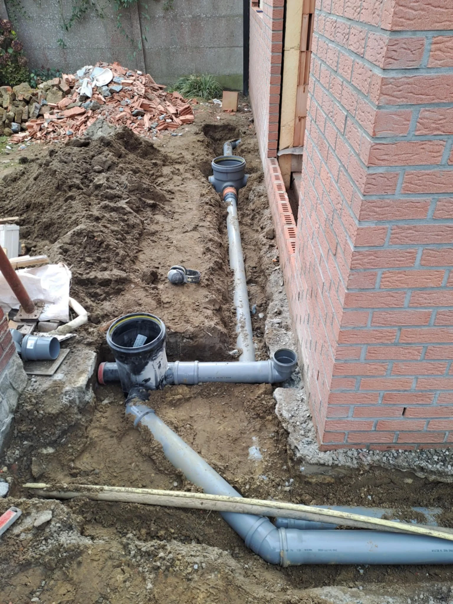 Bodem is gegraven voor installatie van riolering rondom een huis, met zichtbare PVC-buizen en een manhole, en een bakstenen muur van het huis.