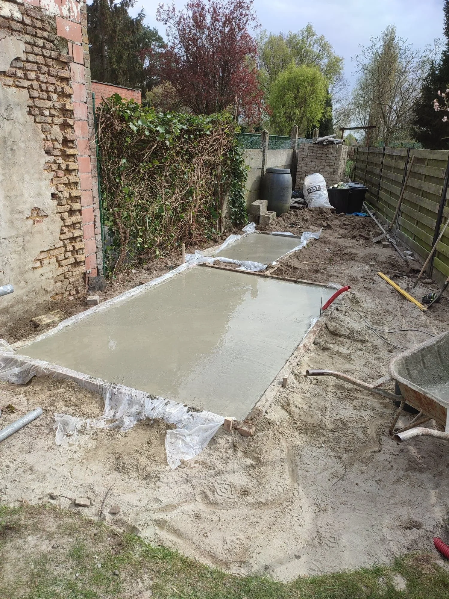 Bouwplaats met vers aangelegde betonnen vloer, omgeven door zand en bouwmaterialen, in een tuin met bomen en houten hek.