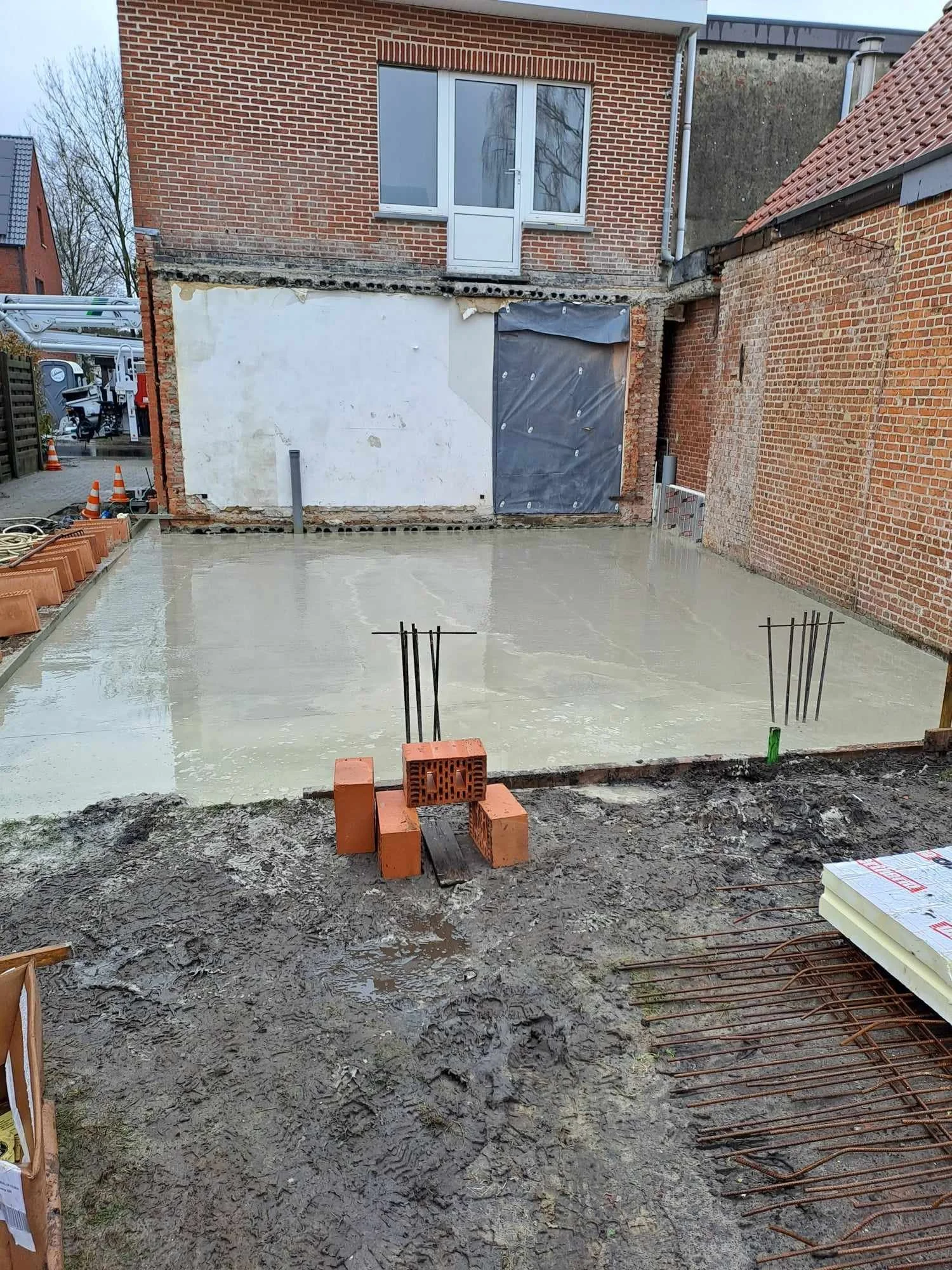 Nieuwe betonnen vloer wordt aangelegd in een achtertuin, met bakstenen en metalen wapening zichtbaar, en een scheiding van muren en een dubbele deur op de achtergrond.