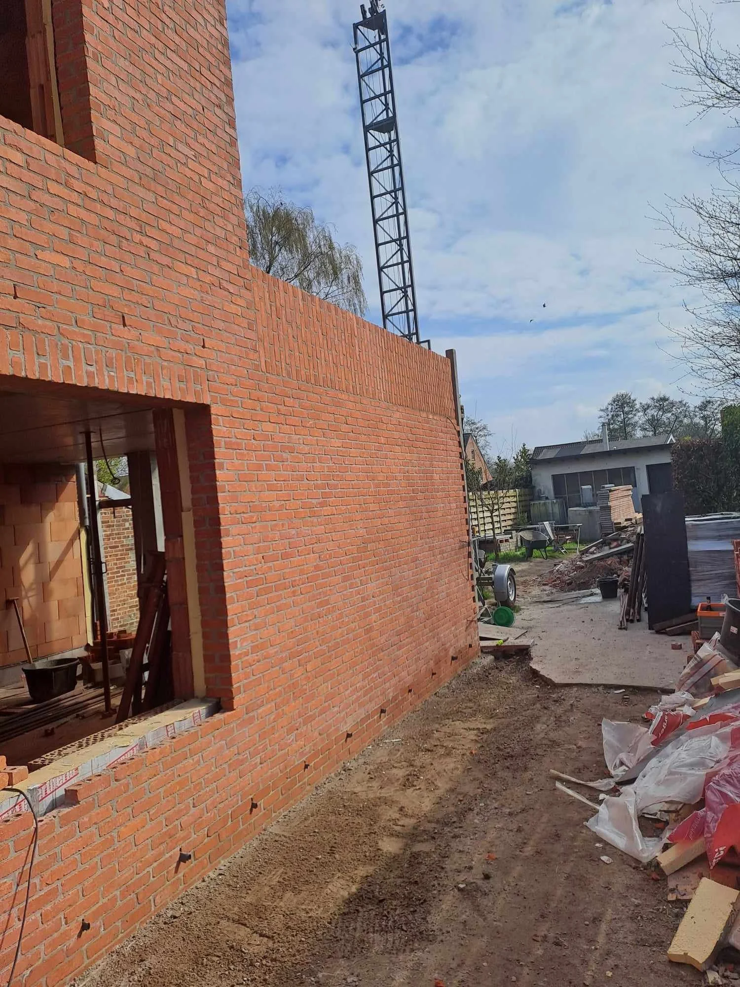 Bouwplaats met een rode bakstenen muur en een open raam, bouwmaterialen en gereedschap op de grond, een telescoopmast op de achtergrond, en een zandpad naast de muur.
