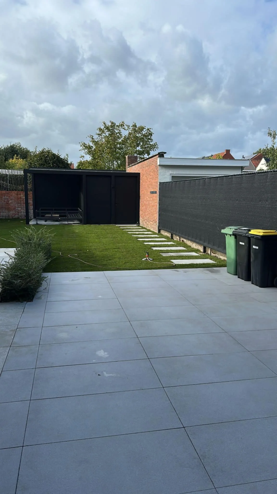 Uitzicht vanaf een terras met grote grijze tegels over een tuin met gazon, plantenbakken en een tuinhek, met een moderne schutting, afvalcontainers, en een gedeeltelijk overdekte zithoek op de achtergrond onder een bewolkte hemel.