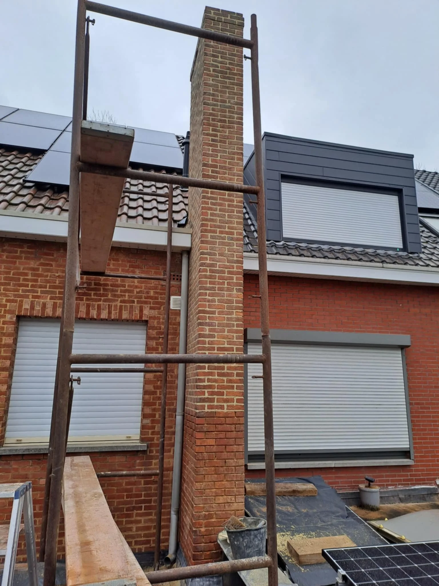Bouwsteiger voor werkzaamheden aan een schoorsteen van een huis met zonnepanelen op het dak en gesloten rolluiken over ramen.