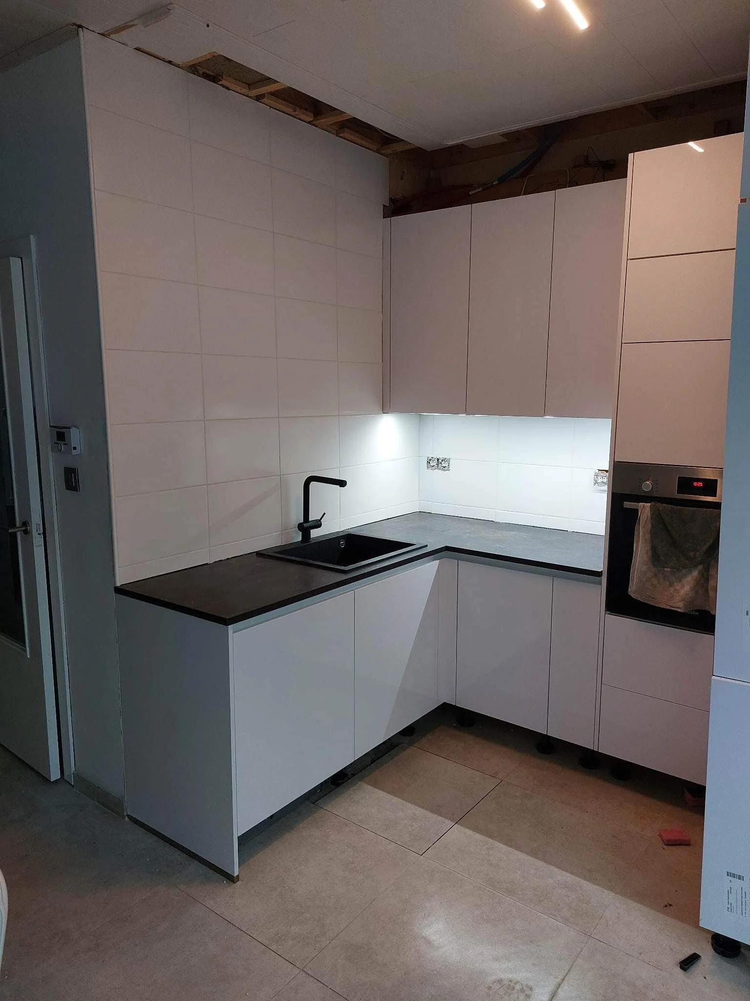 Moderne keuken met witte kasten, zwart fornuis, en inbouwoven, nog in aanbouw met opvallend plafond en tilwerkzaamheden boven de kasten.