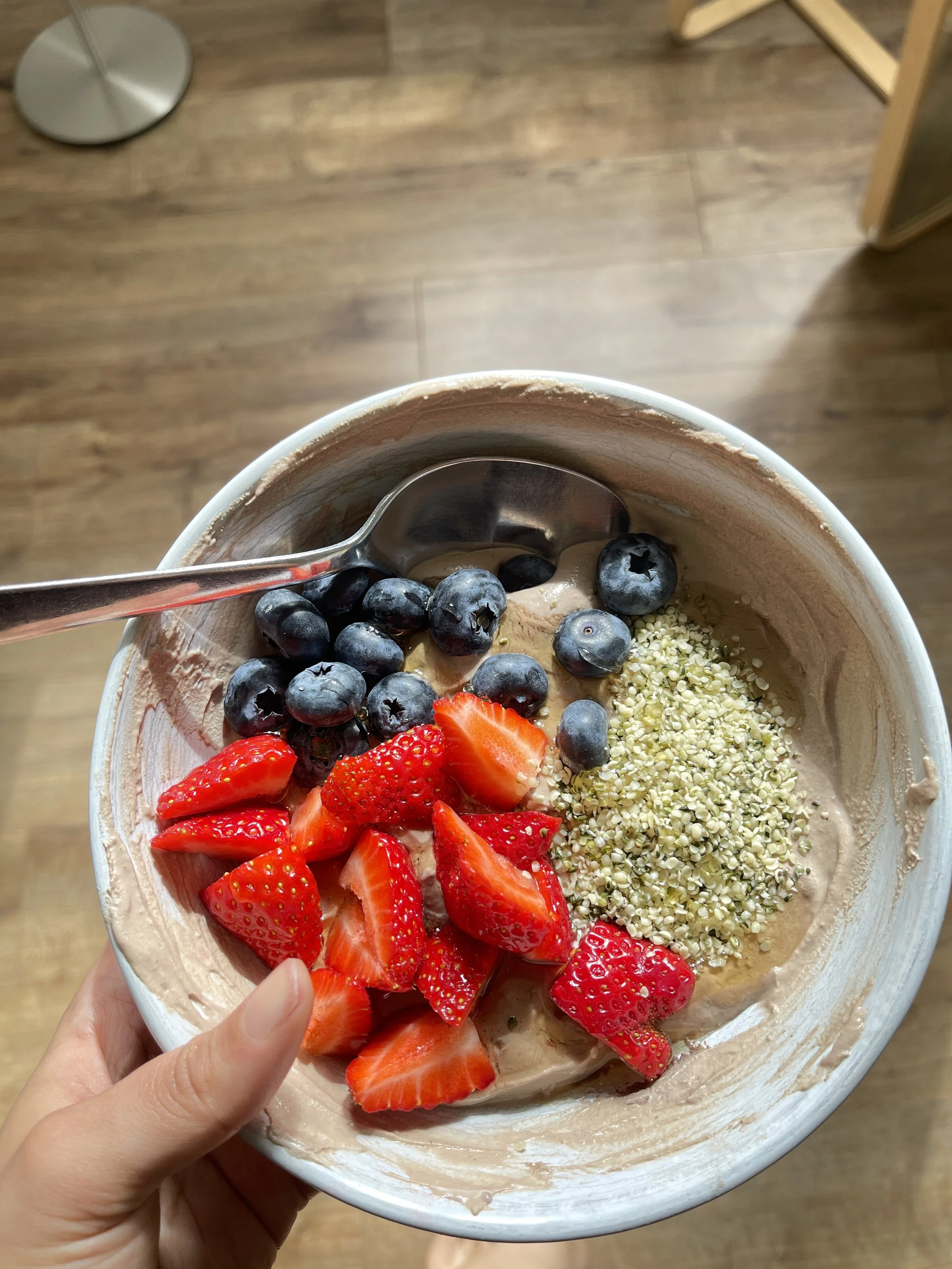 A bowl of chocolate protein yoghurt topped with fresh blueberries, sliced strawberries, and hemp seeds, with a spoon inside.