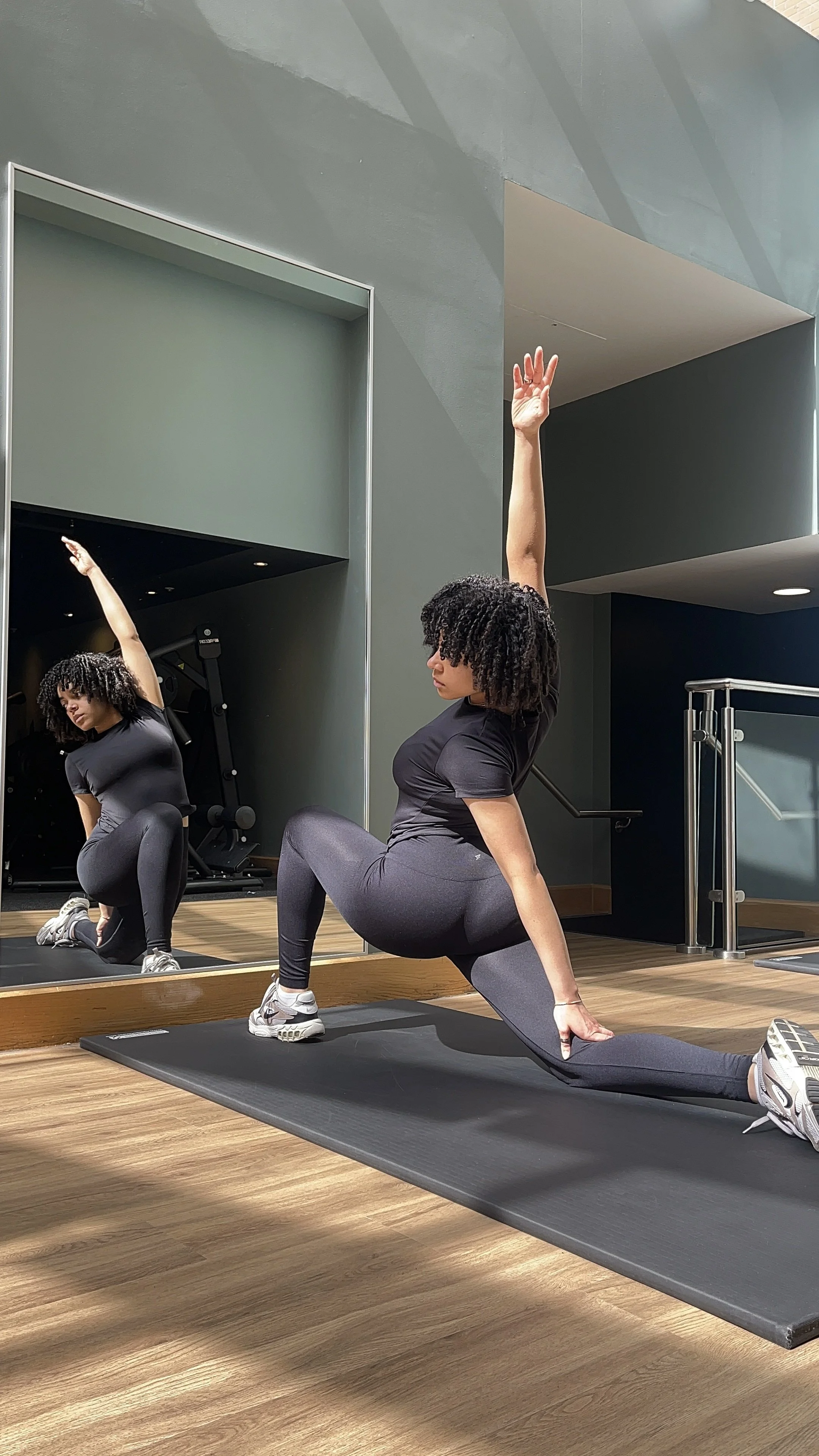 A woman in workout clothes performs a stretching exercise on a black yoga mat in front of a mirror at a gym. She is in a lunge position with one arm raised, looking at her reflection.