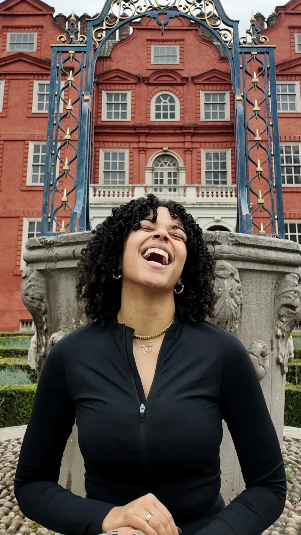 A woman with curly hair laughing in front of a historic red-brick building with ornate architectural details and a decorative iron gate.