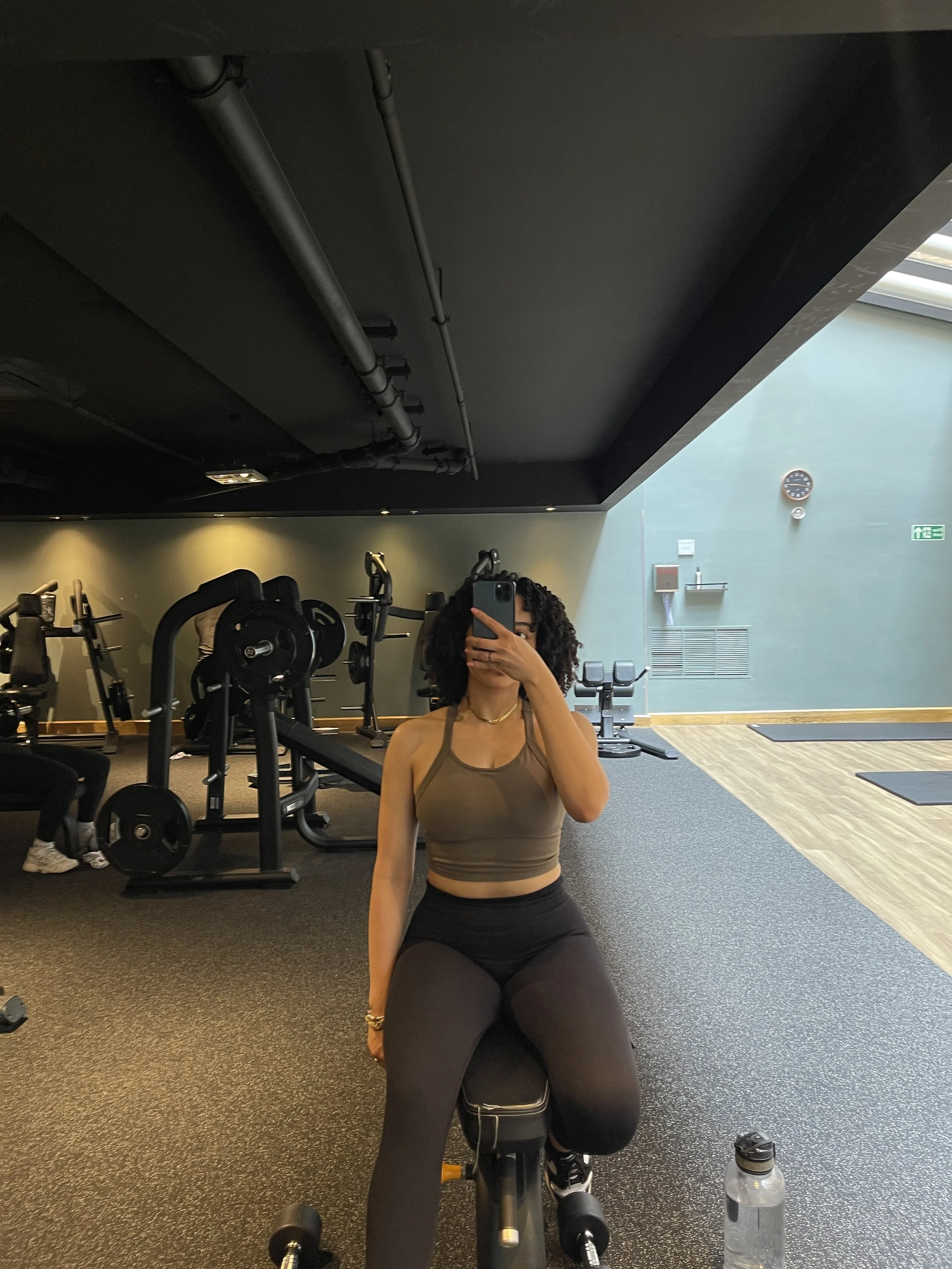 A woman in workout attire taking a selfie in a gym mirror, sitting on an exercise bench with gym equipment in the background.