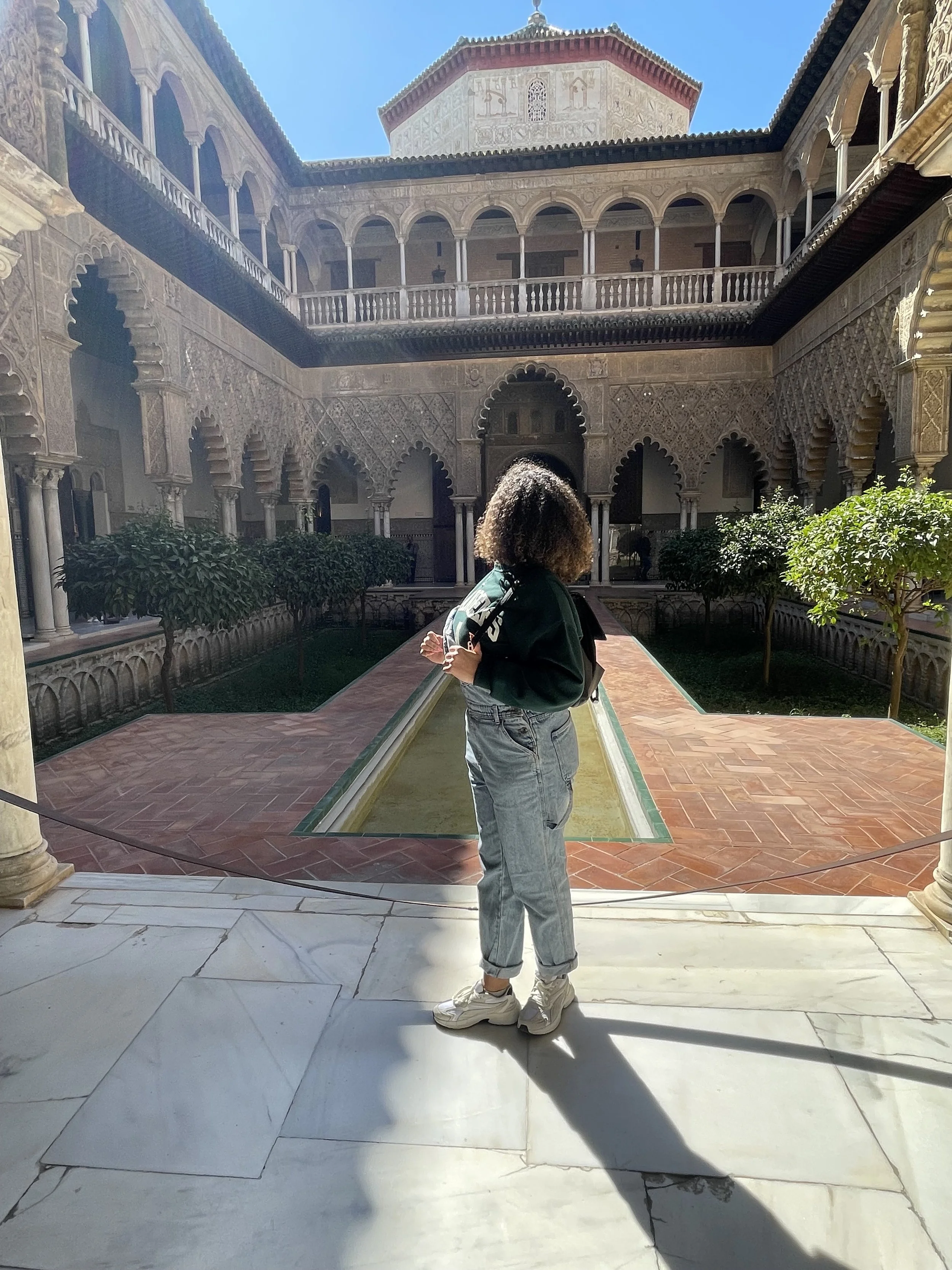 A person with curly hair wearing a dark jacket and jeans standing in a courtyard of an ornate building with arches, columns, and intricate architectural details, with a small garden and a fountain in the center.