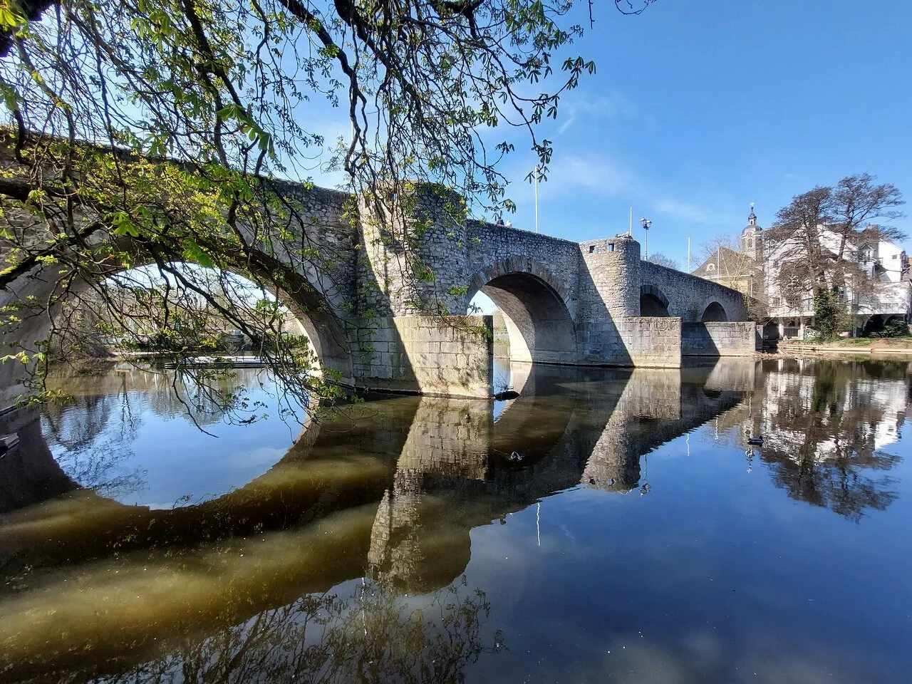Een oude stenen brug over een rivier met reflectie, omgeven door bomen en gebouwen in de achtergrond, heldere blauwe hemel.