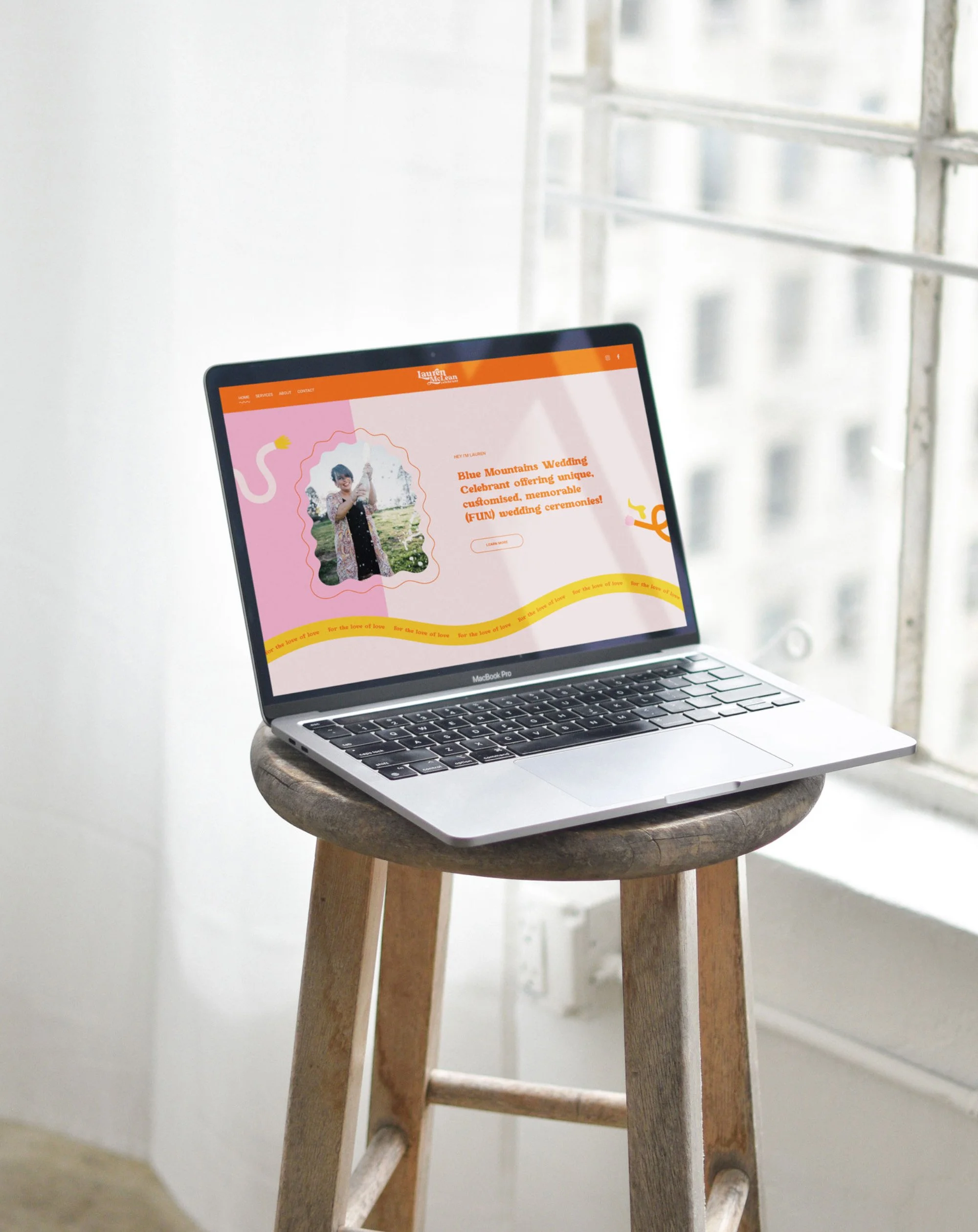 A MacBook Pro laptop on a wooden stool displaying a website for Blue Mountains Wedding Celebrant, with a pink and orange colorful background and a photo of a woman holding a child.