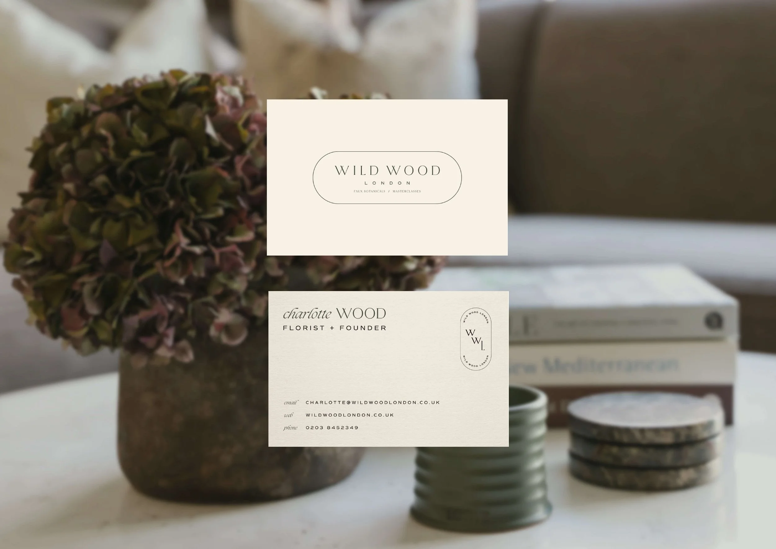 Business cards for Wild Wood London, a florist and founder Charlotte Wood, placed on a table with a vase of dried hydrangeas, a stack of coasters, and books in the background.