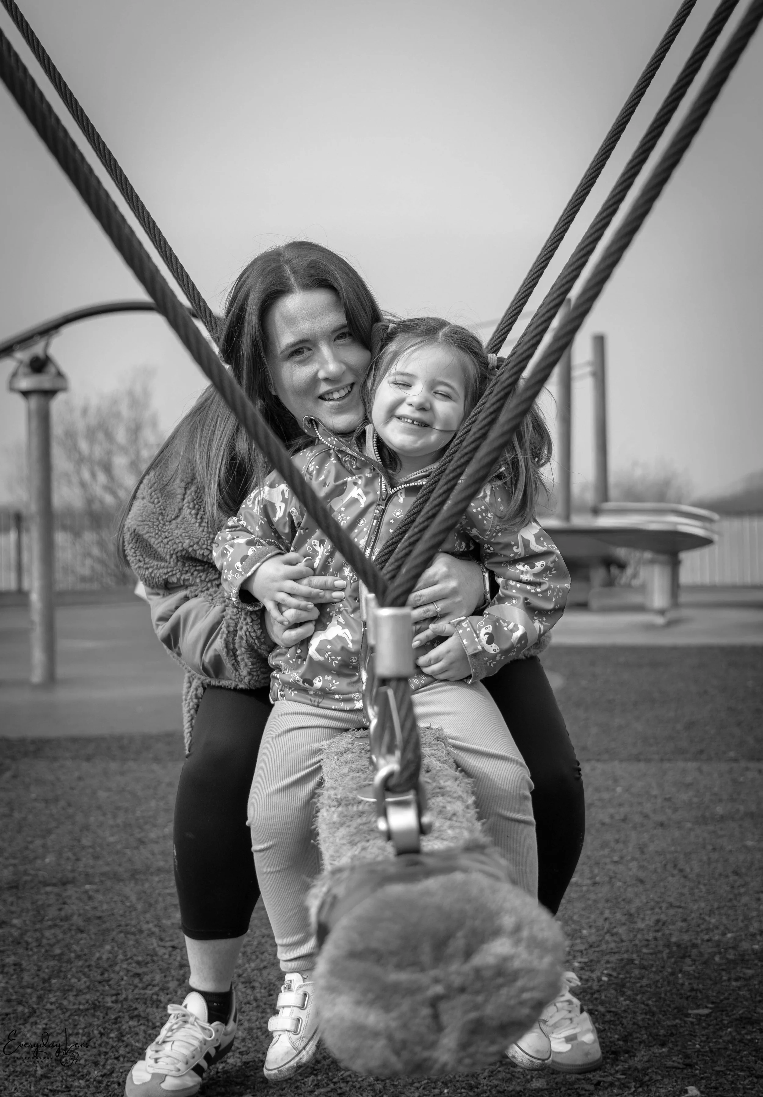 A woman and a young girl smiling and hugging while sitting on a swing at a playground.