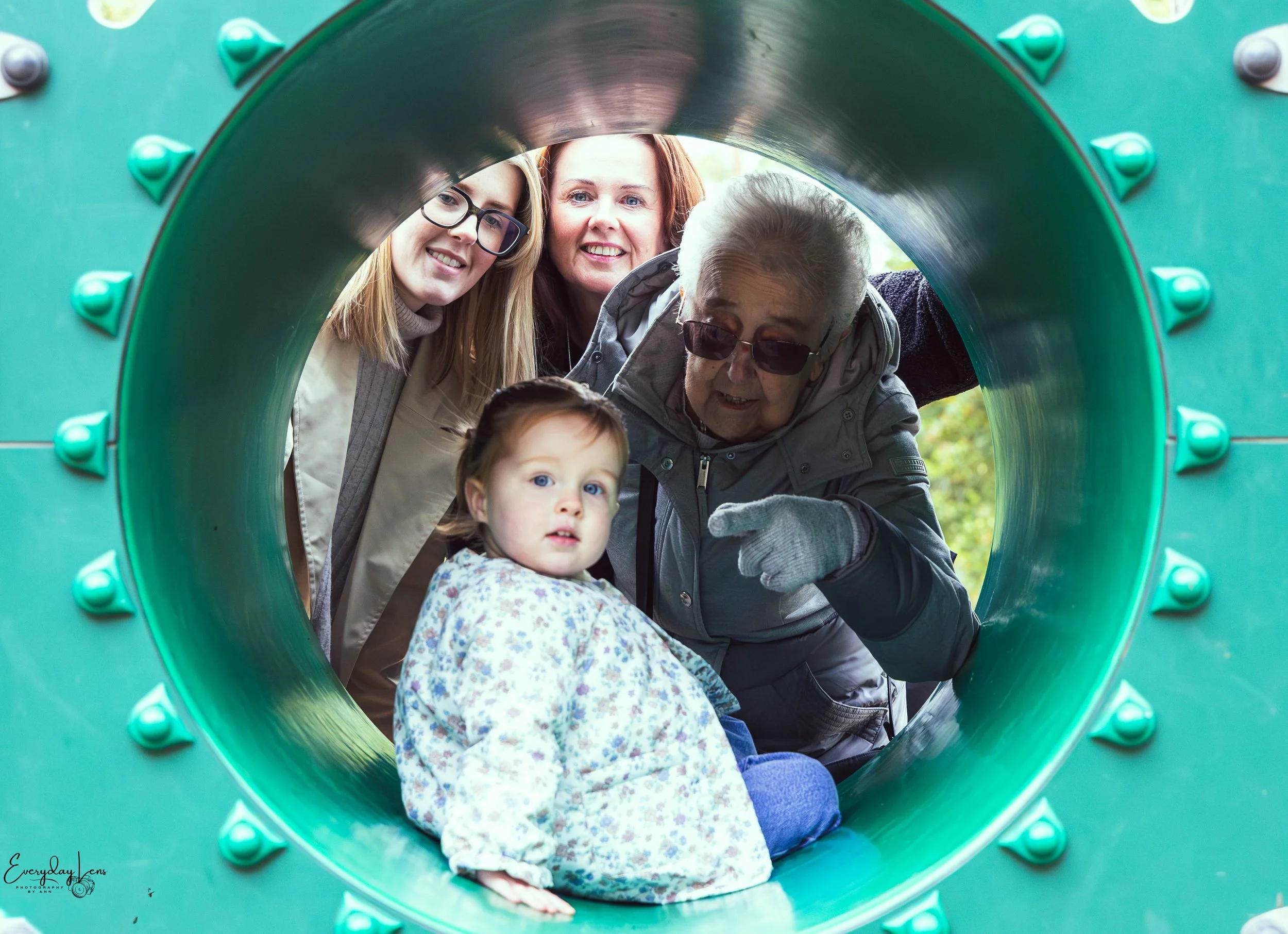 Group of four people, two women, a girl, and a woman with glasses, looking through a green cylindrical playground tunnel, with trees in the background.