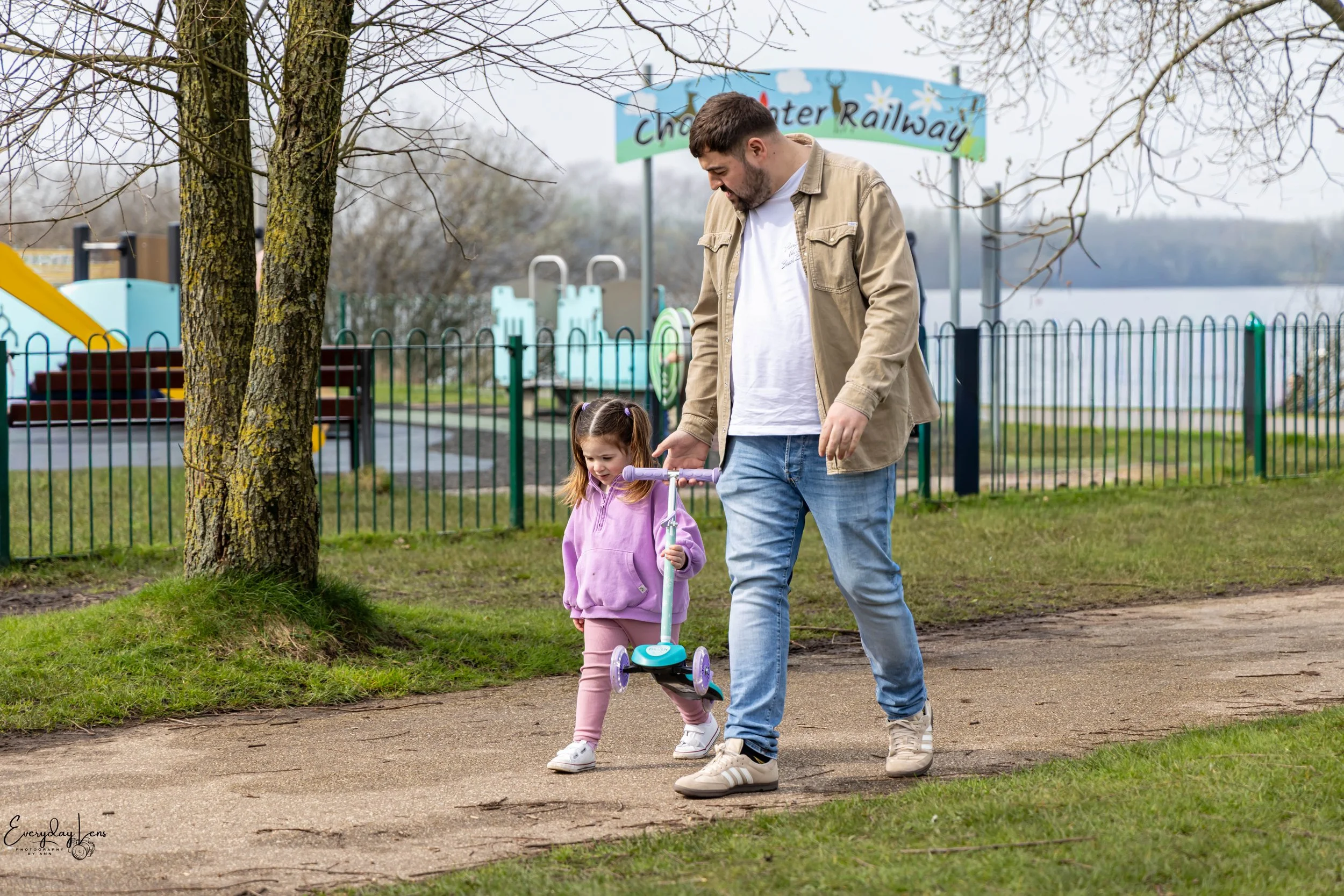 A father and daughter walking on a dirt path near a playground in a park, with trees and a fence in the background. The girl is holding a purple scooter.