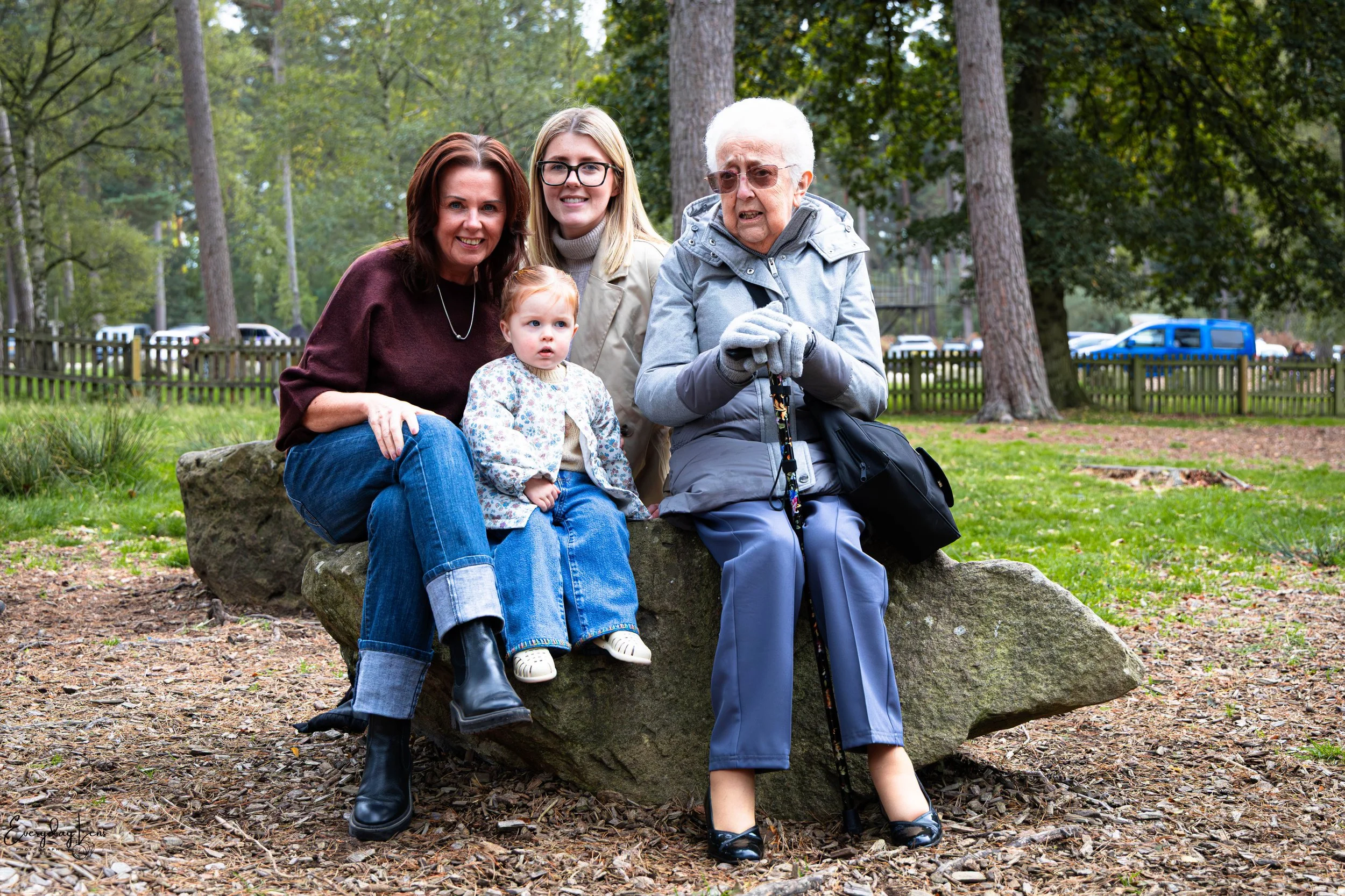 Three women and a young girl sitting on a large rock in a park with trees and a wooden fence in the background.