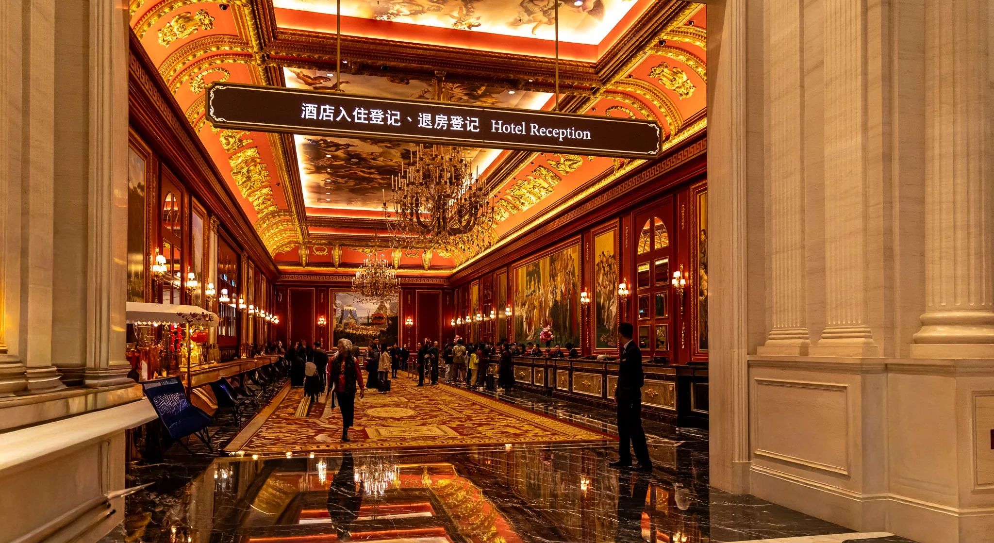 Interior of a luxurious hotel lobby with high ornate ceiling, chandeliers, large paintings on the walls, and a reception desk with people waiting. A sign hanging from the ceiling reads 'Hotel Reception' in both Chinese and English.