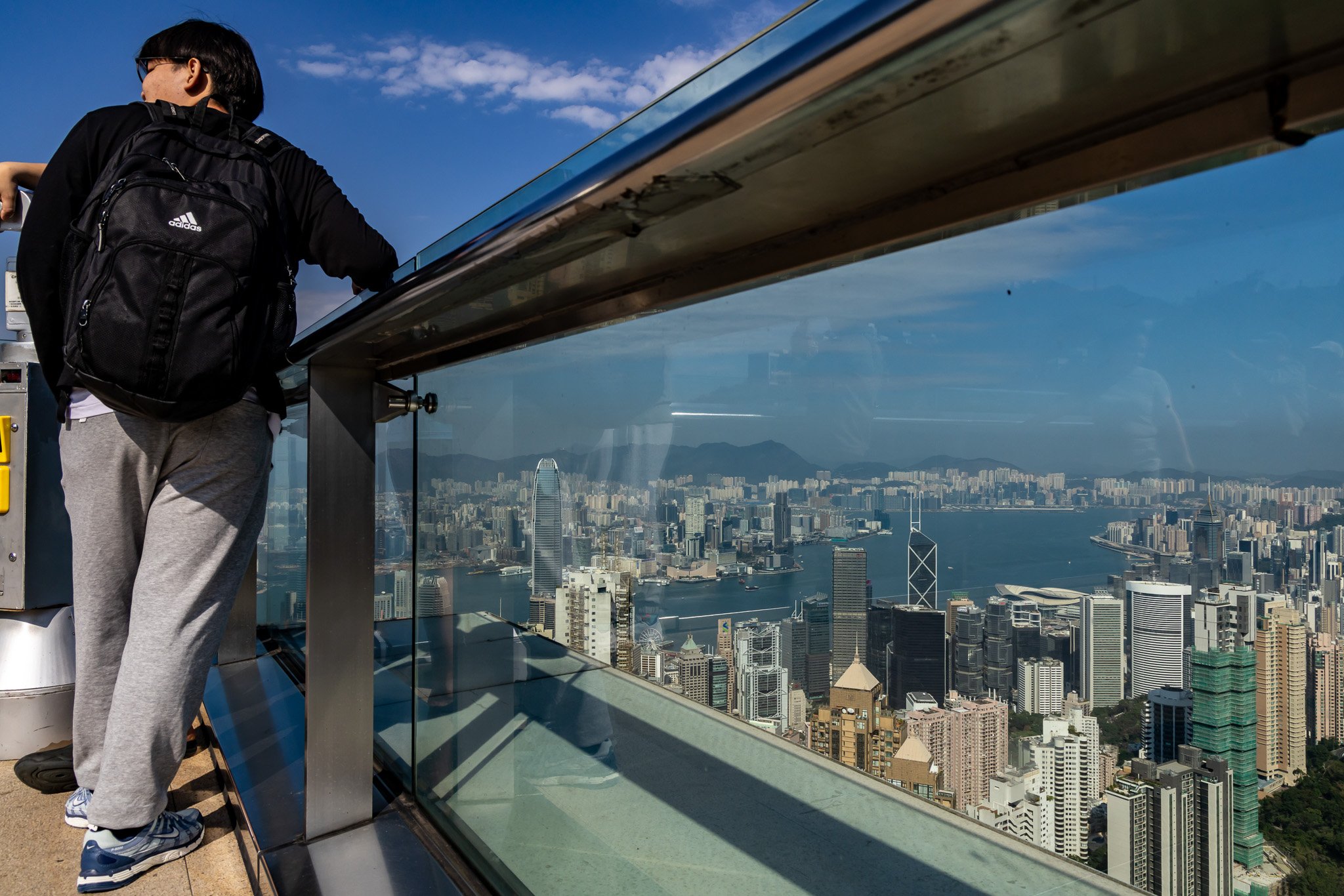 A person with a backpack looking over the glass railing of an observation deck at a city skyline, including tall buildings and a body of water, under a partly cloudy sky.