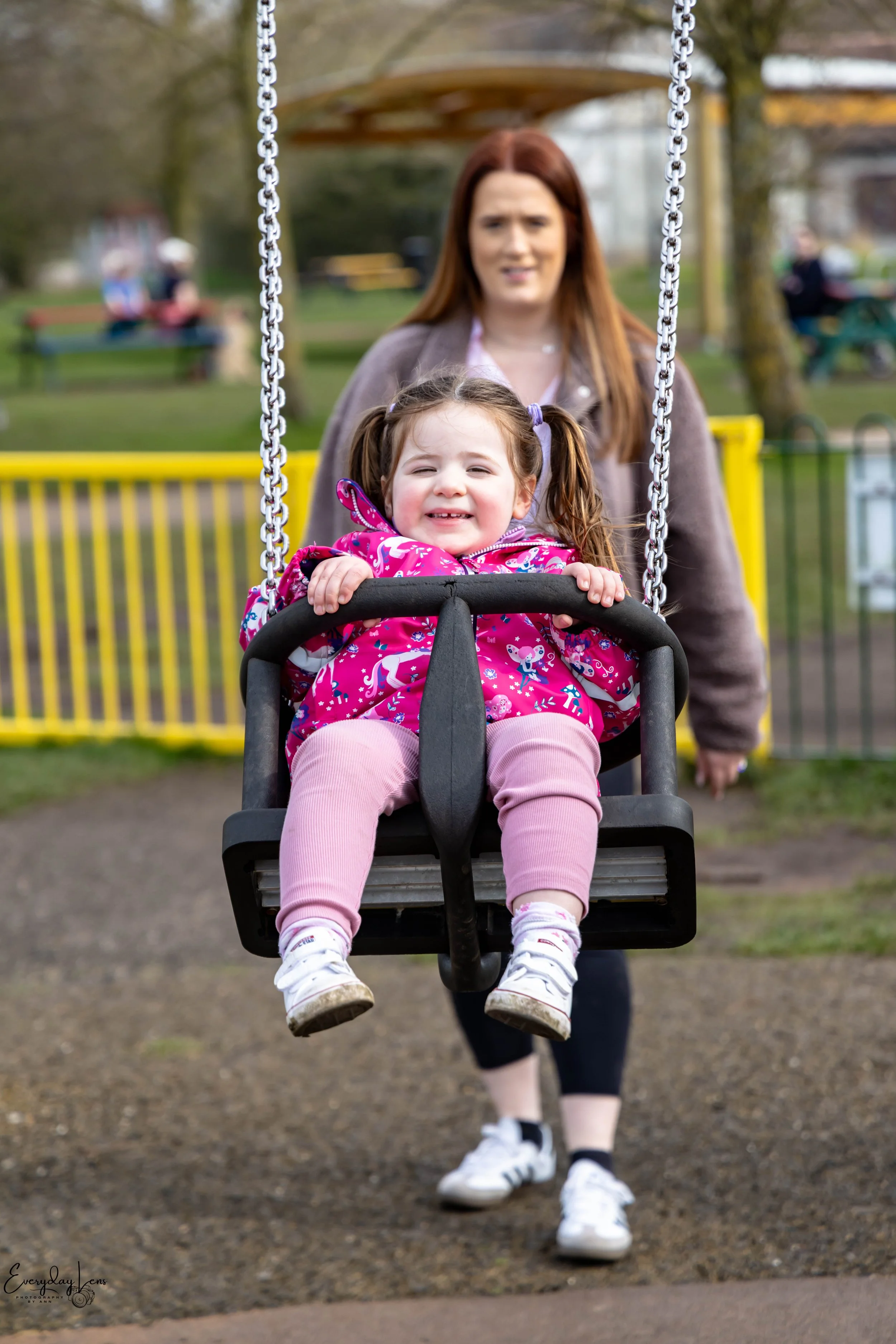 A young girl smiling on a swing with a woman behind her at a park.