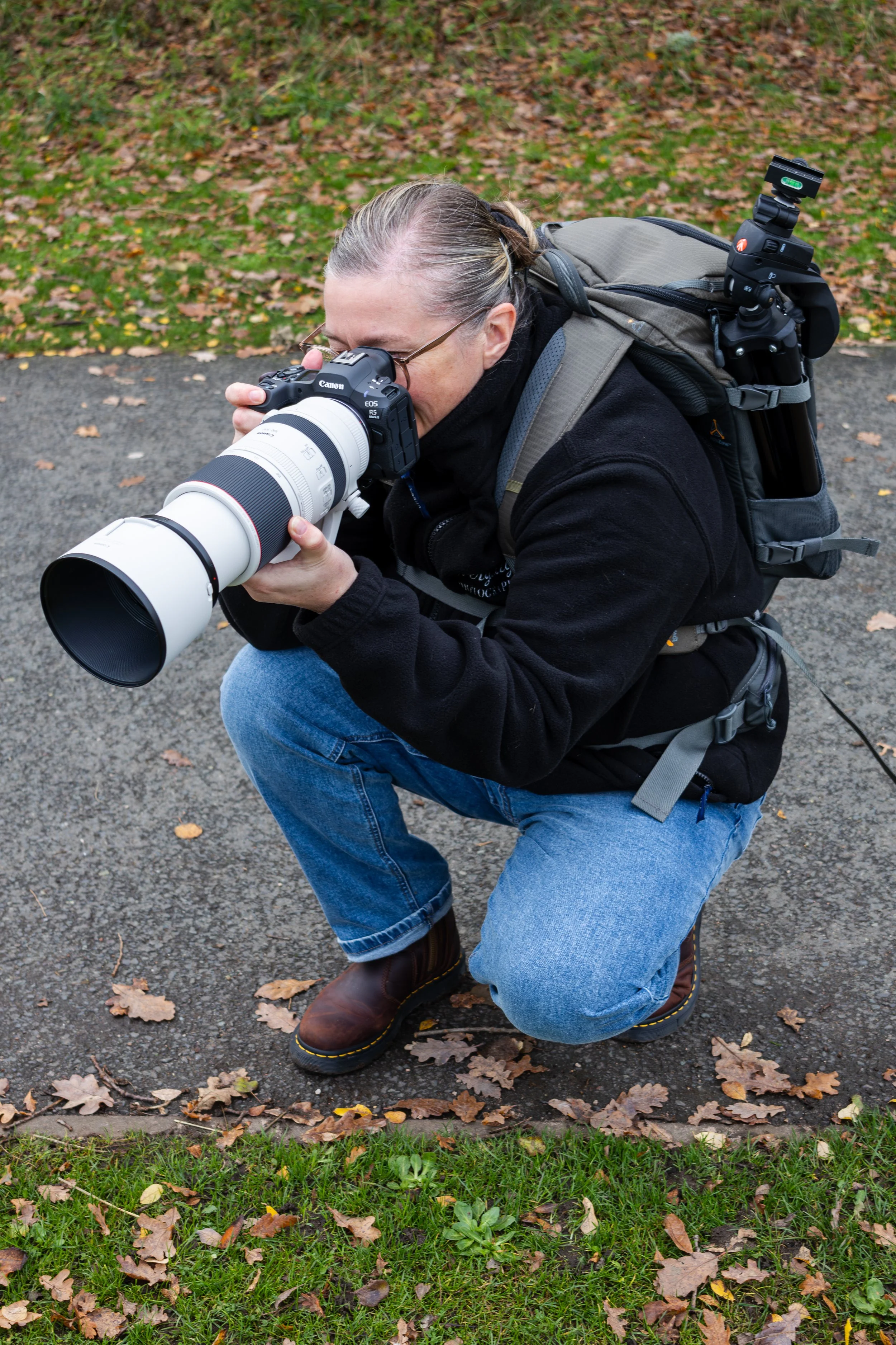Person crouching on the sidewalk taking a photograph with a professional camera, surrounded by autumn leaves.