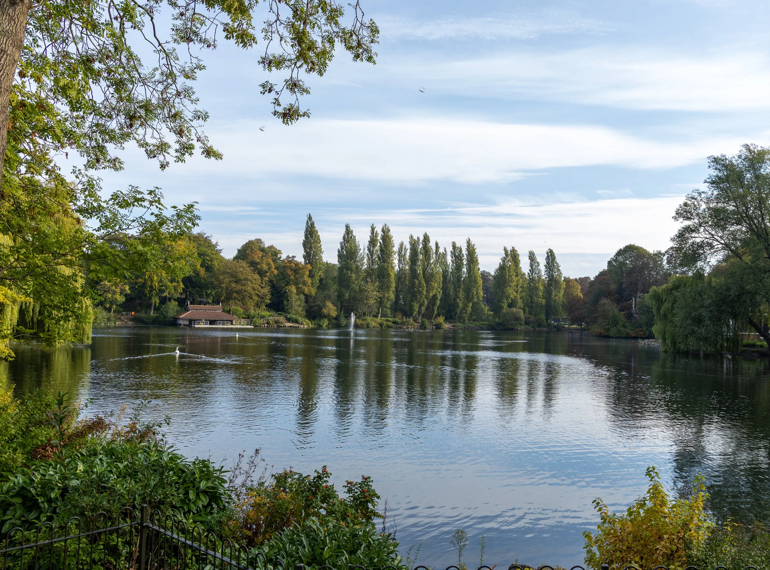 A scenic view of a lake surrounded by trees and greenery, with a small building and fountain visible on the far shore, under a partly cloudy sky.