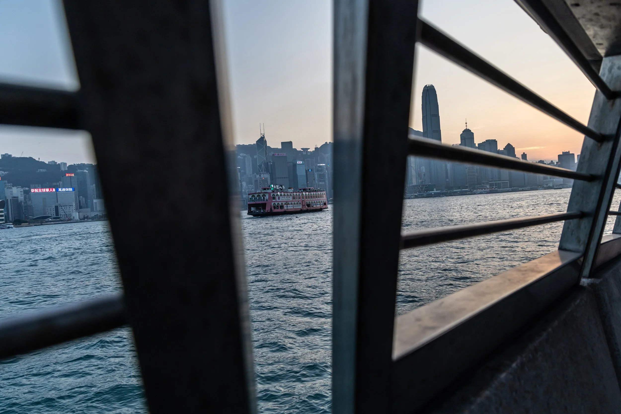 View of Hong Kong skyline through metal railings, including a pink ferry on the water during sunset.