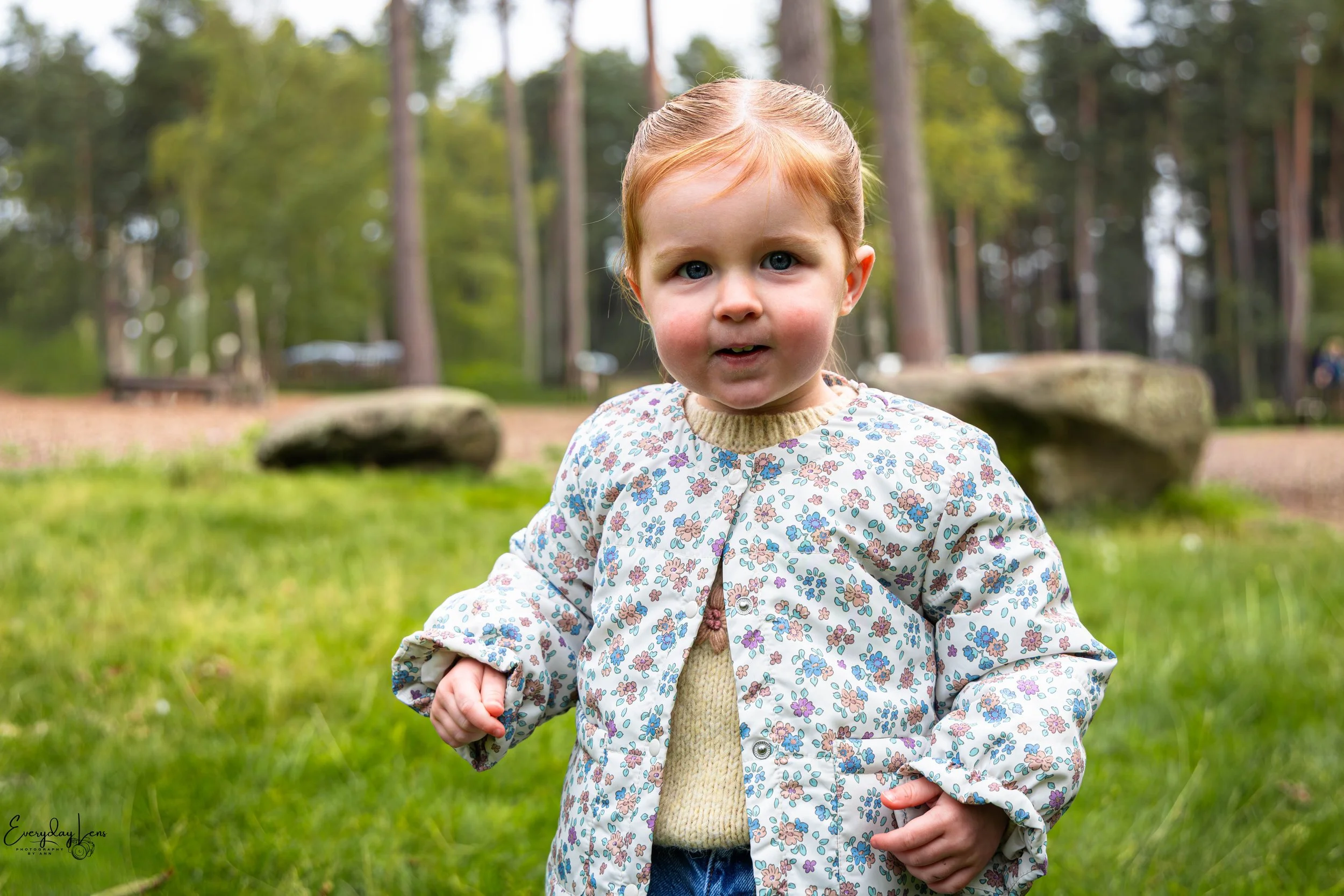 A young girl with red hair, blue eyes, and a floral jacket standing outdoors in a wooded area with trees and large rocks in the background.