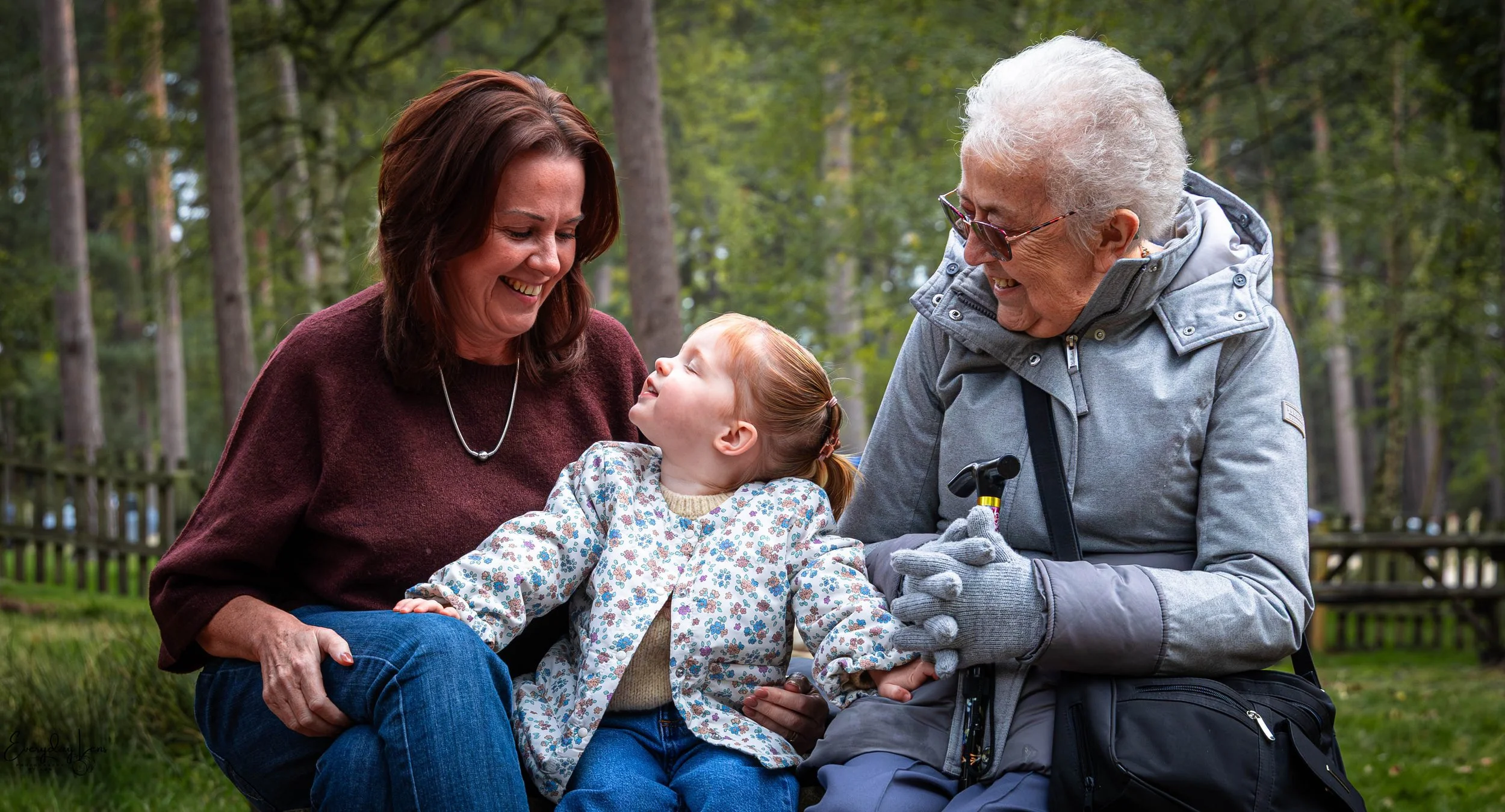 Three females and one young girl sitting outdoors in a wooded area, smiling and enjoying each other's company, with trees and a fence in the background.