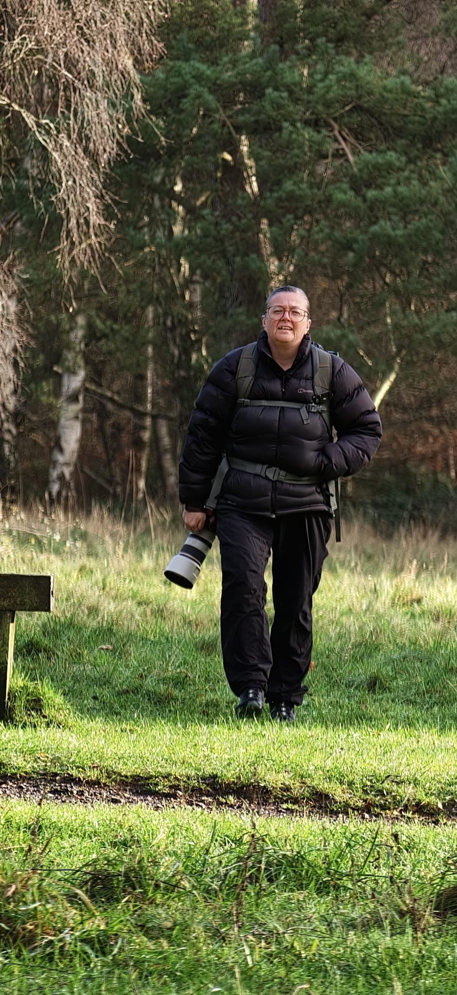 A person with glasses carrying a camera walks through a grassy area with trees in the background.