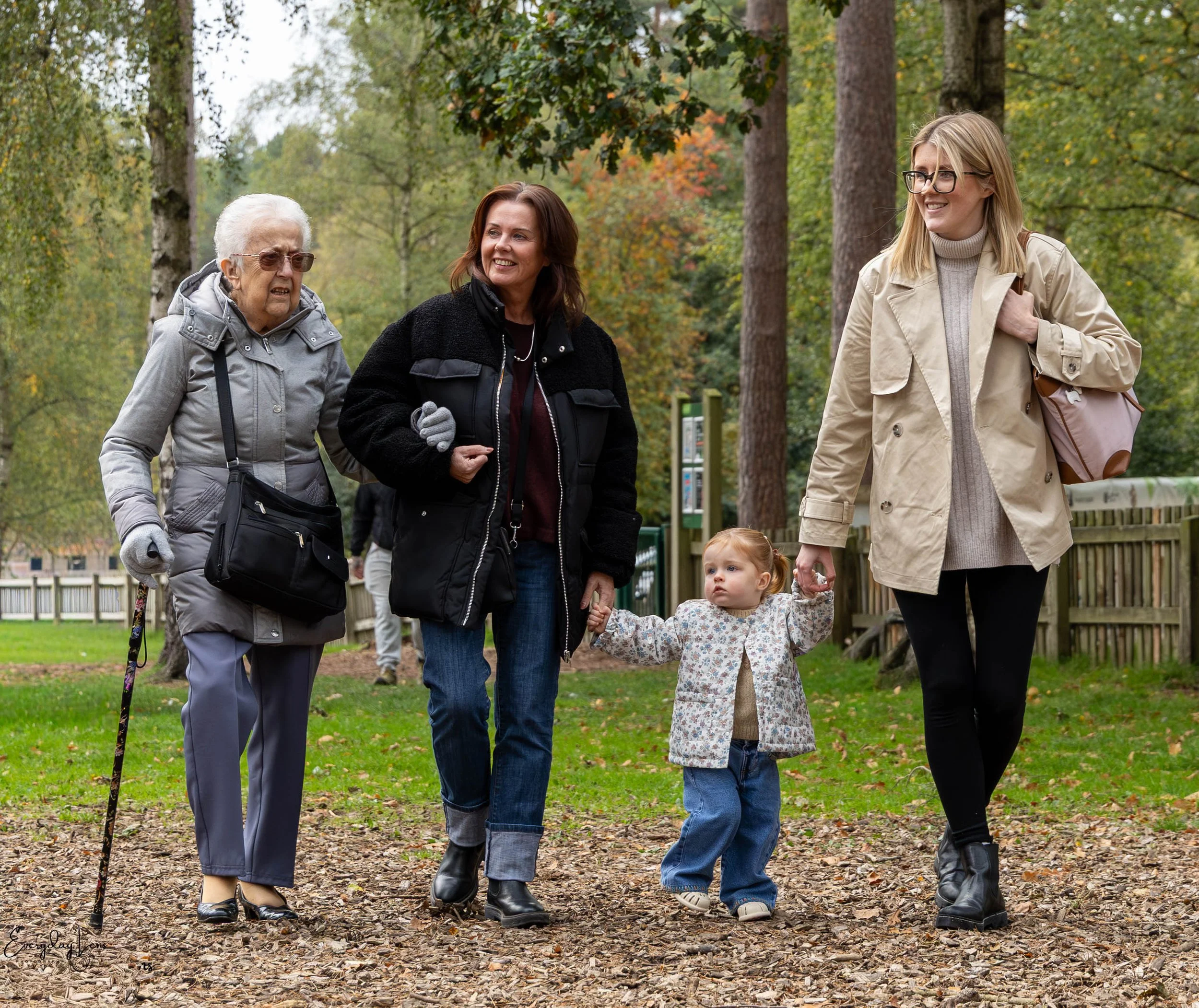 Three women and a young girl walking in a park, holding hands, surrounded by trees with fall foliage.