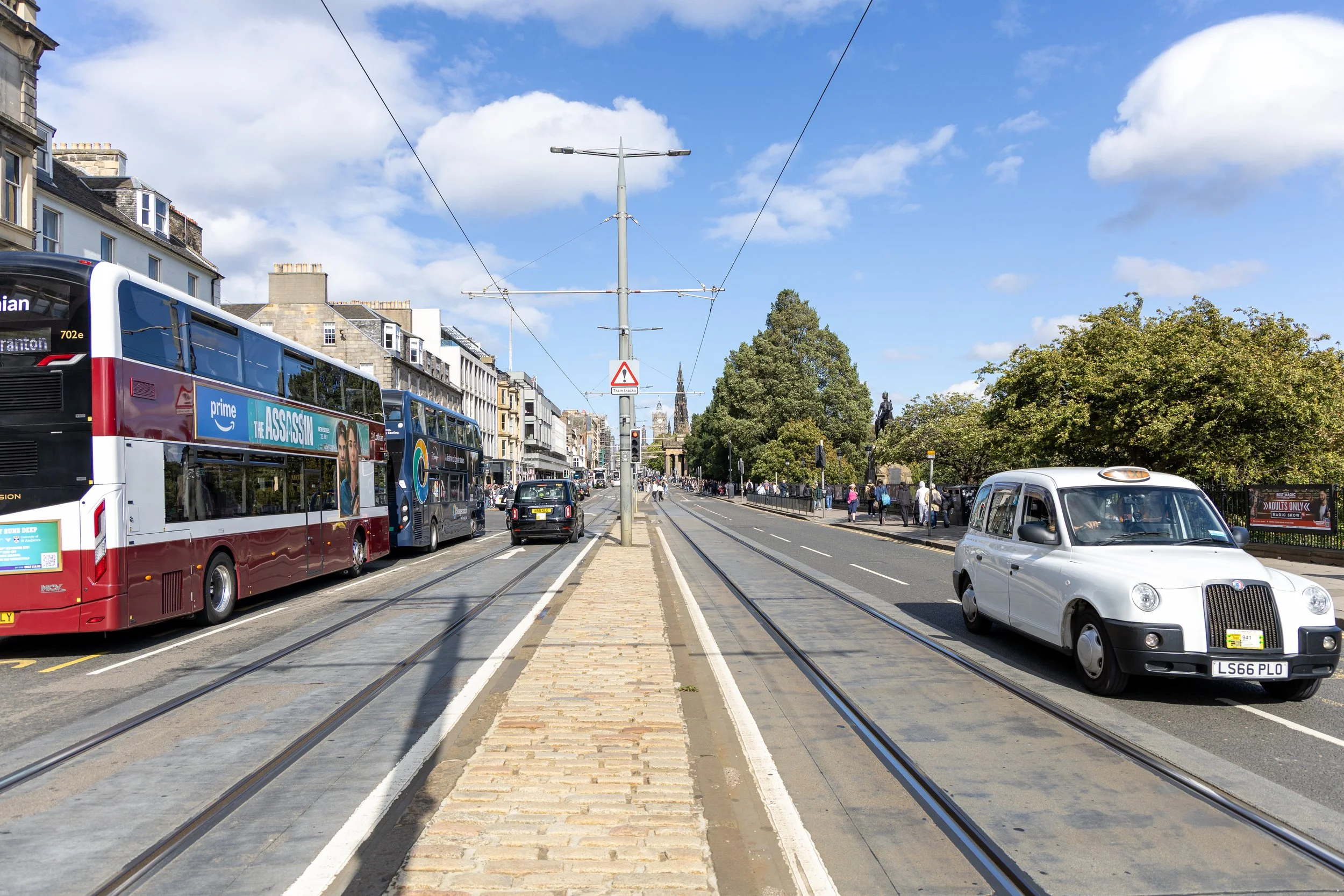 Street scene with a double-decker bus, cars, and pedestrians, tram tracks running down the middle, and buildings lining the street under a blue sky with clouds.