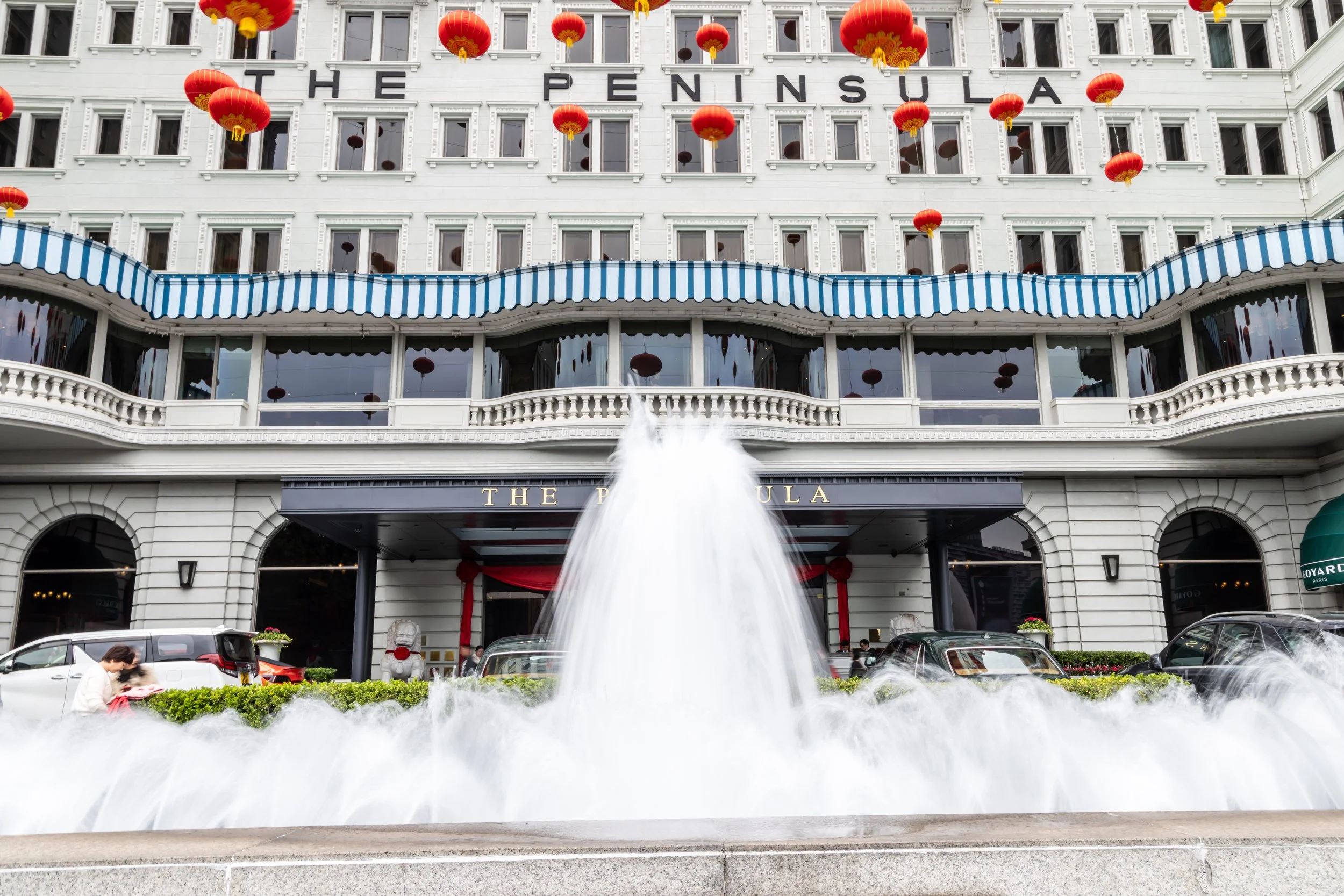 Exterior of The Peninsula hotel decorated with red lanterns, featuring a water fountain in front, cars parked along the street, and a woman taking a photo, indicating a celebration or event.
