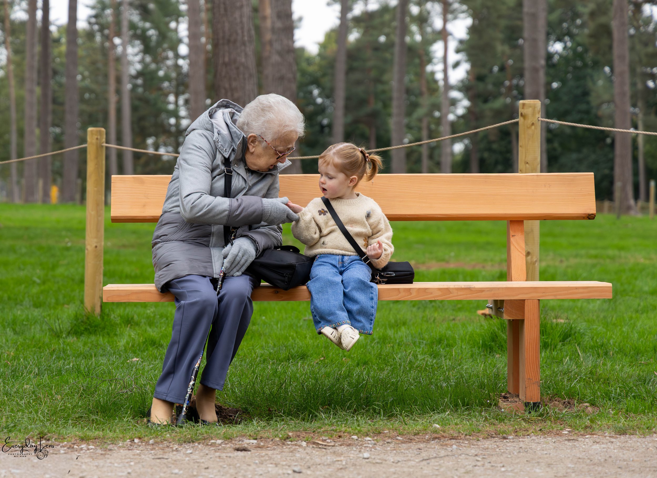 An elderly woman and a young girl sitting on a wooden park bench in a forested area, engaging in conversation.
