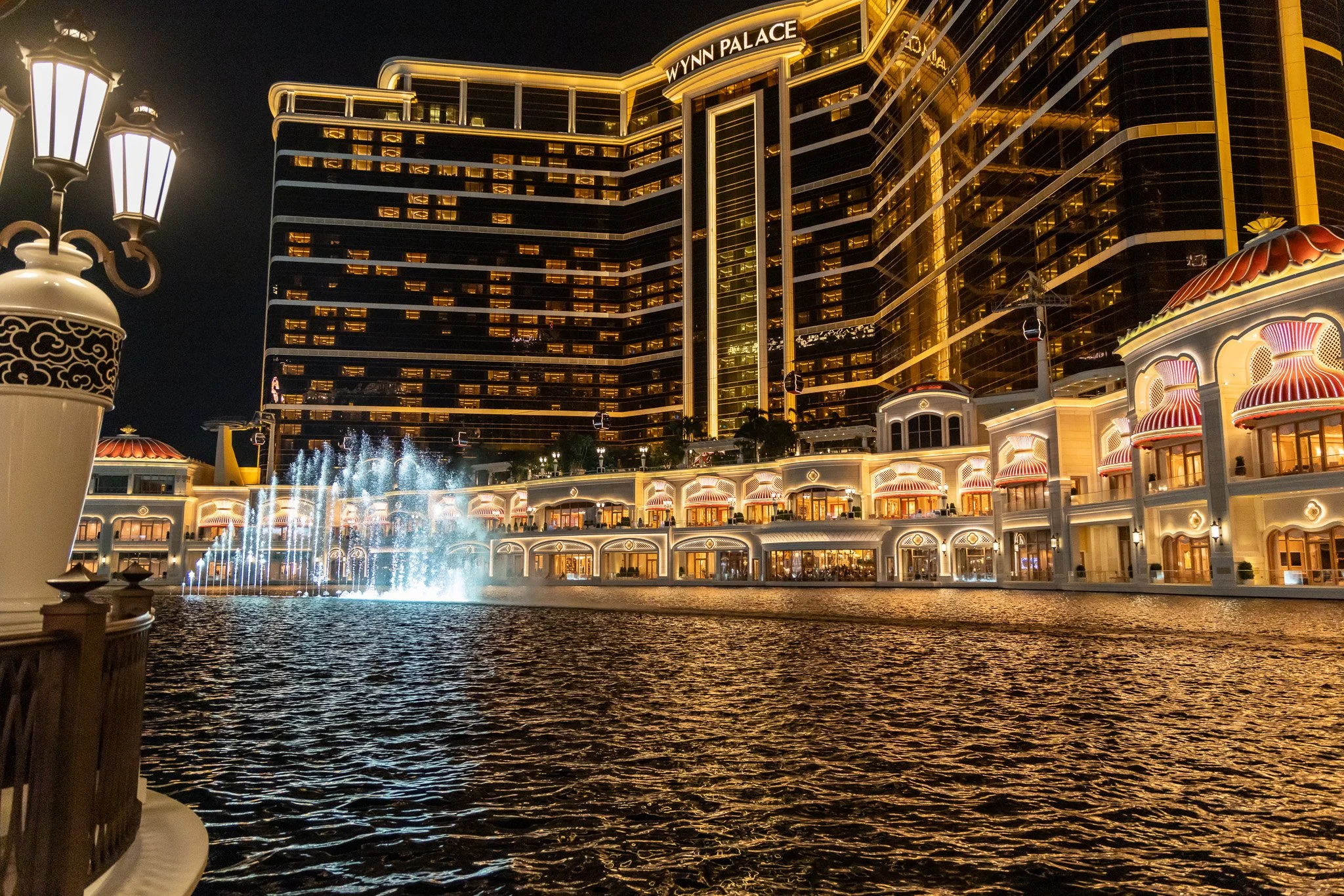 Night view of Wynn Palace hotel in Macau with illuminated building, water fountain, and reflections on water.