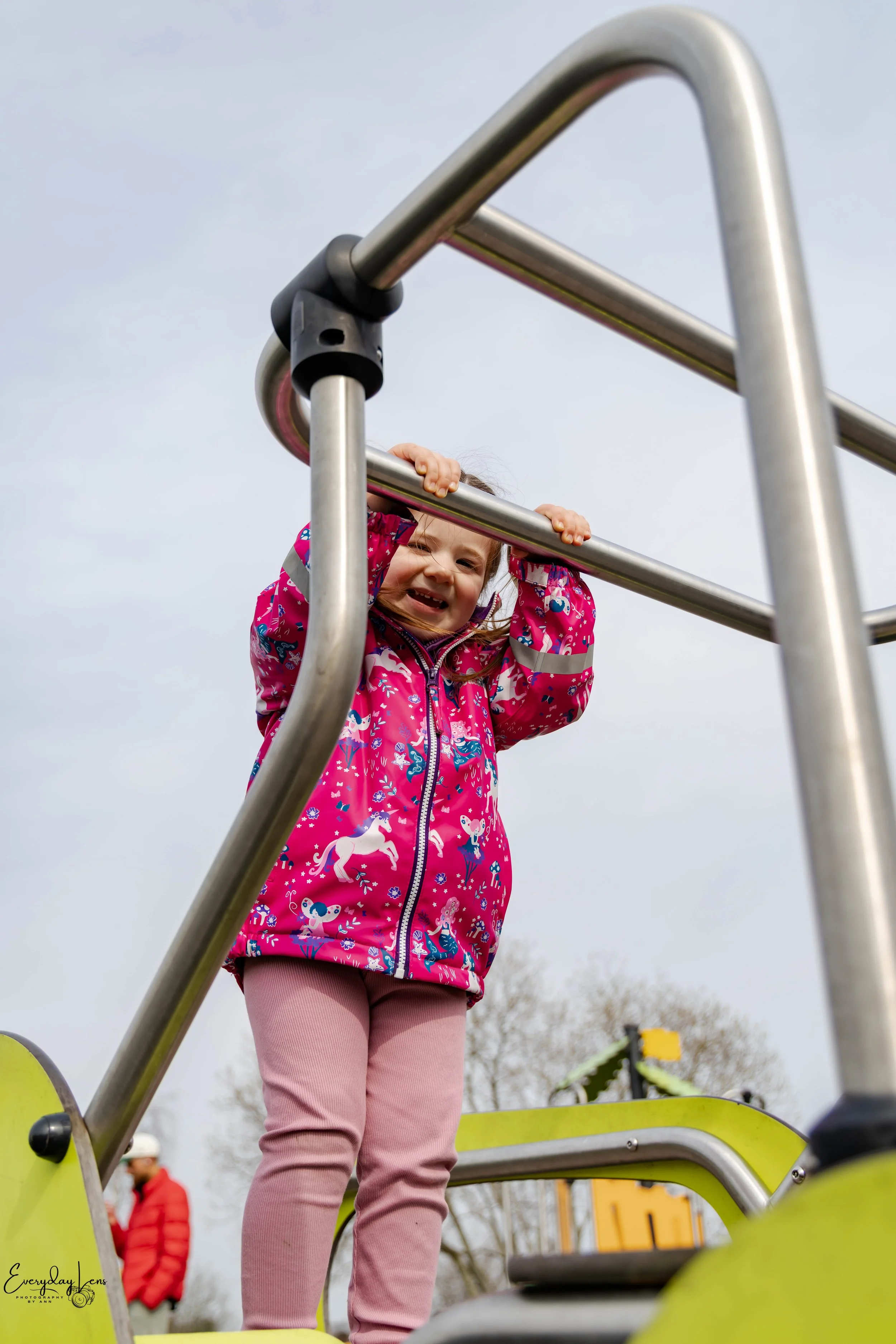 A young girl with a pink jacket and pink pants playing on playground equipment, holding onto the metal bars of a climbing structure, smiling and looking down.
