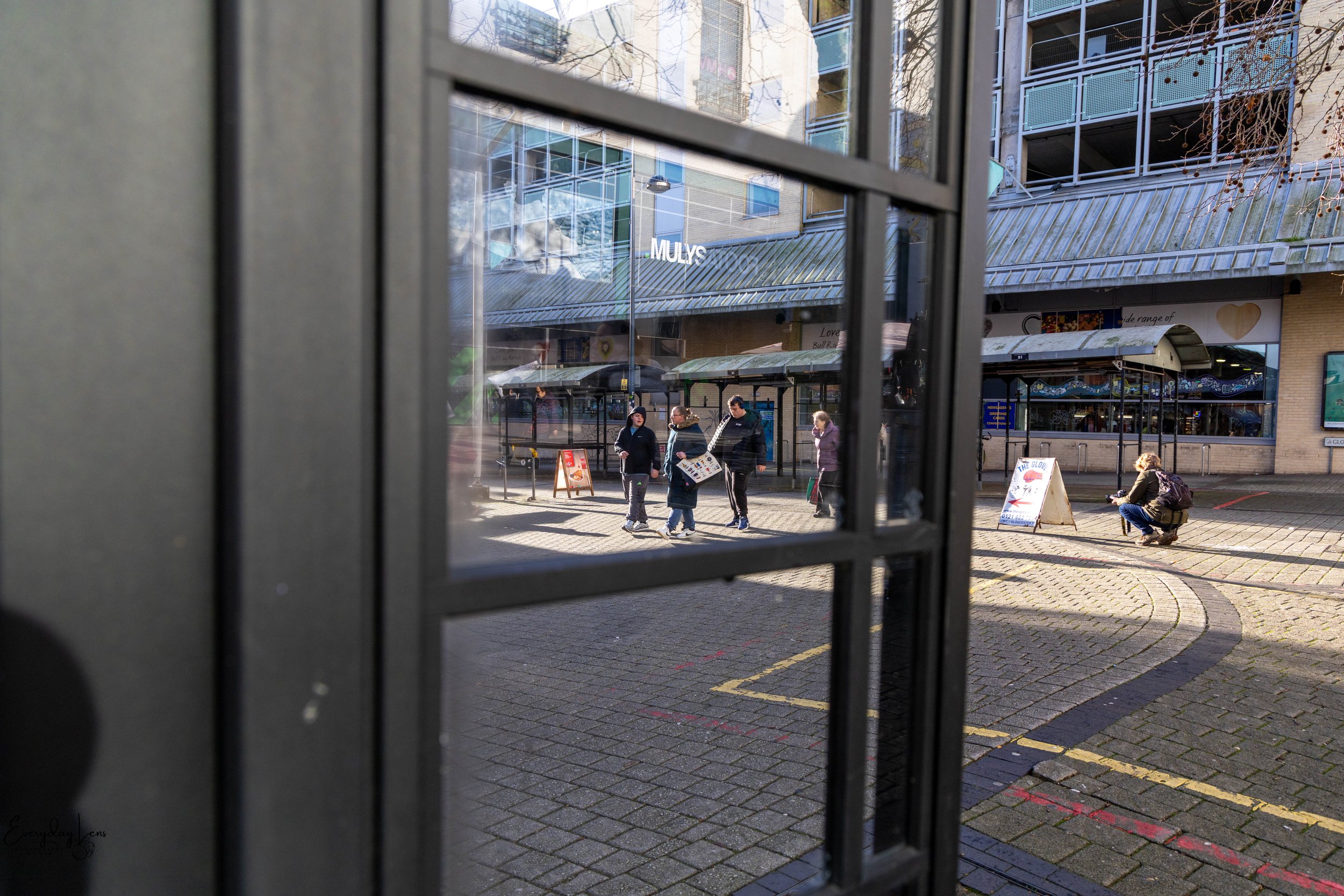 People walking and sitting outside a shopping area seen through a glass window frame.