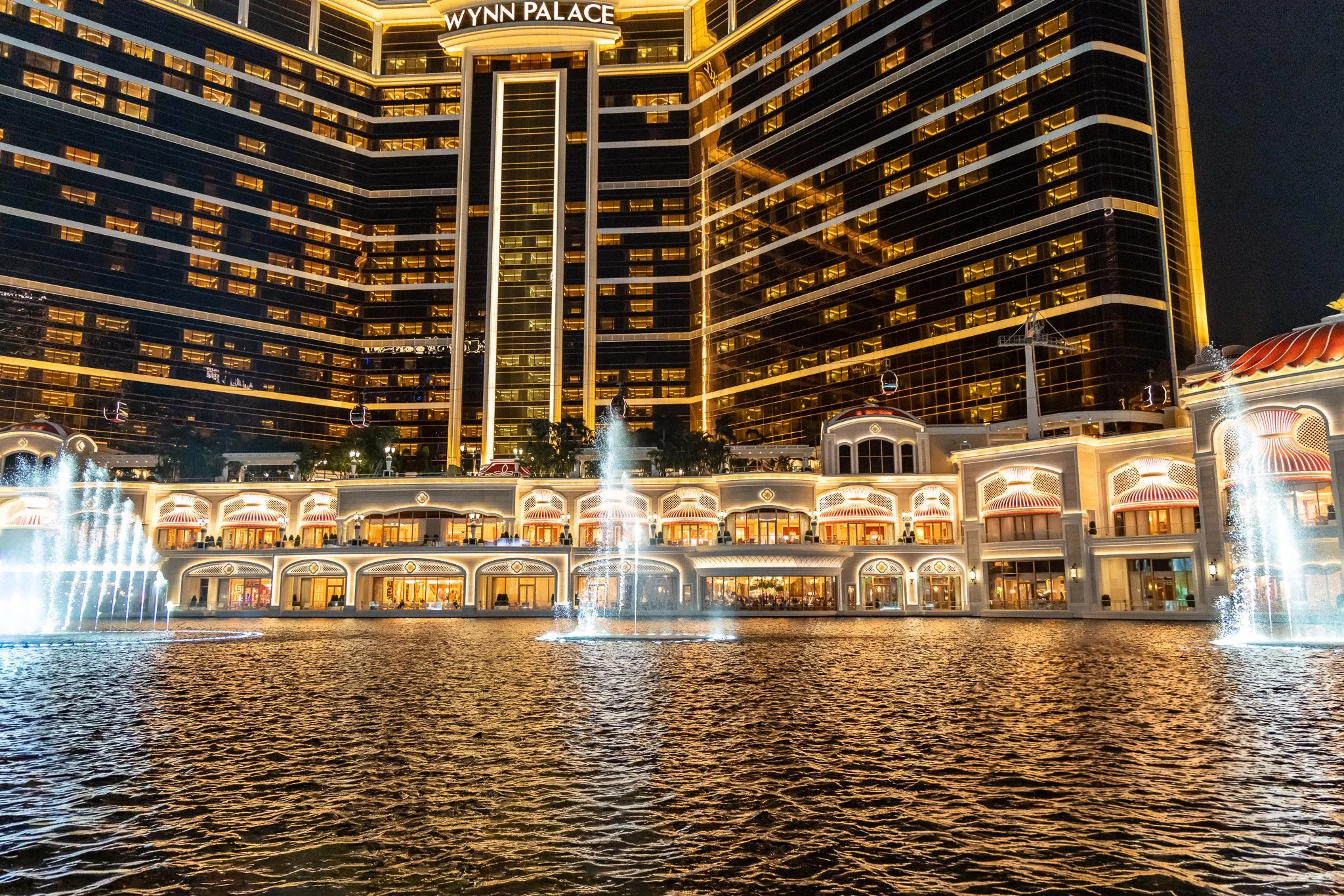 Night view of Wynn Palace hotel with illuminated facade, reflecting on water fountain, and a large modern glass hotel building in the background.