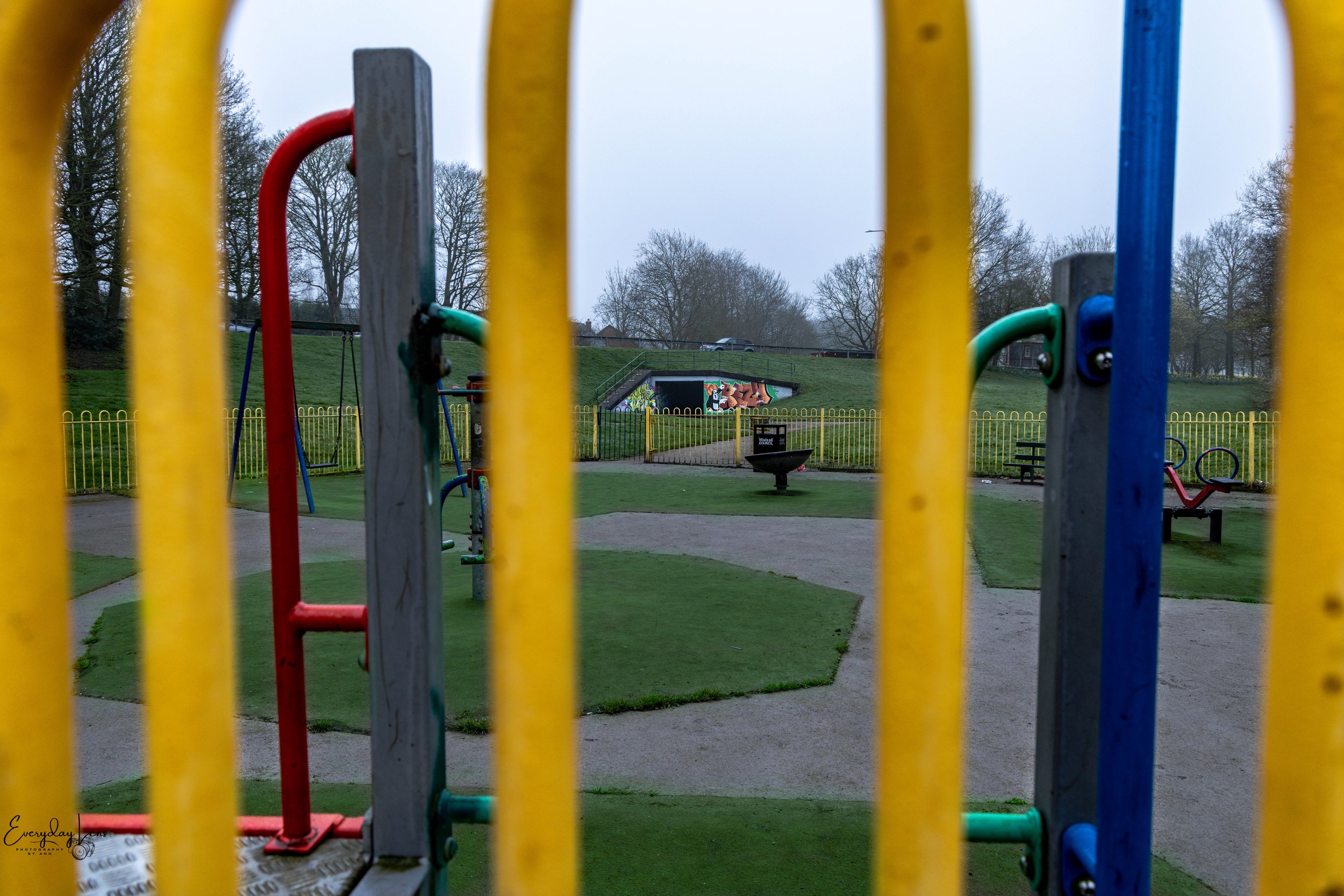 View of a playground area through colorful yellow, red, blue, and green bars, with a grassy hill and trees in the background, and a mural on a tunnel.