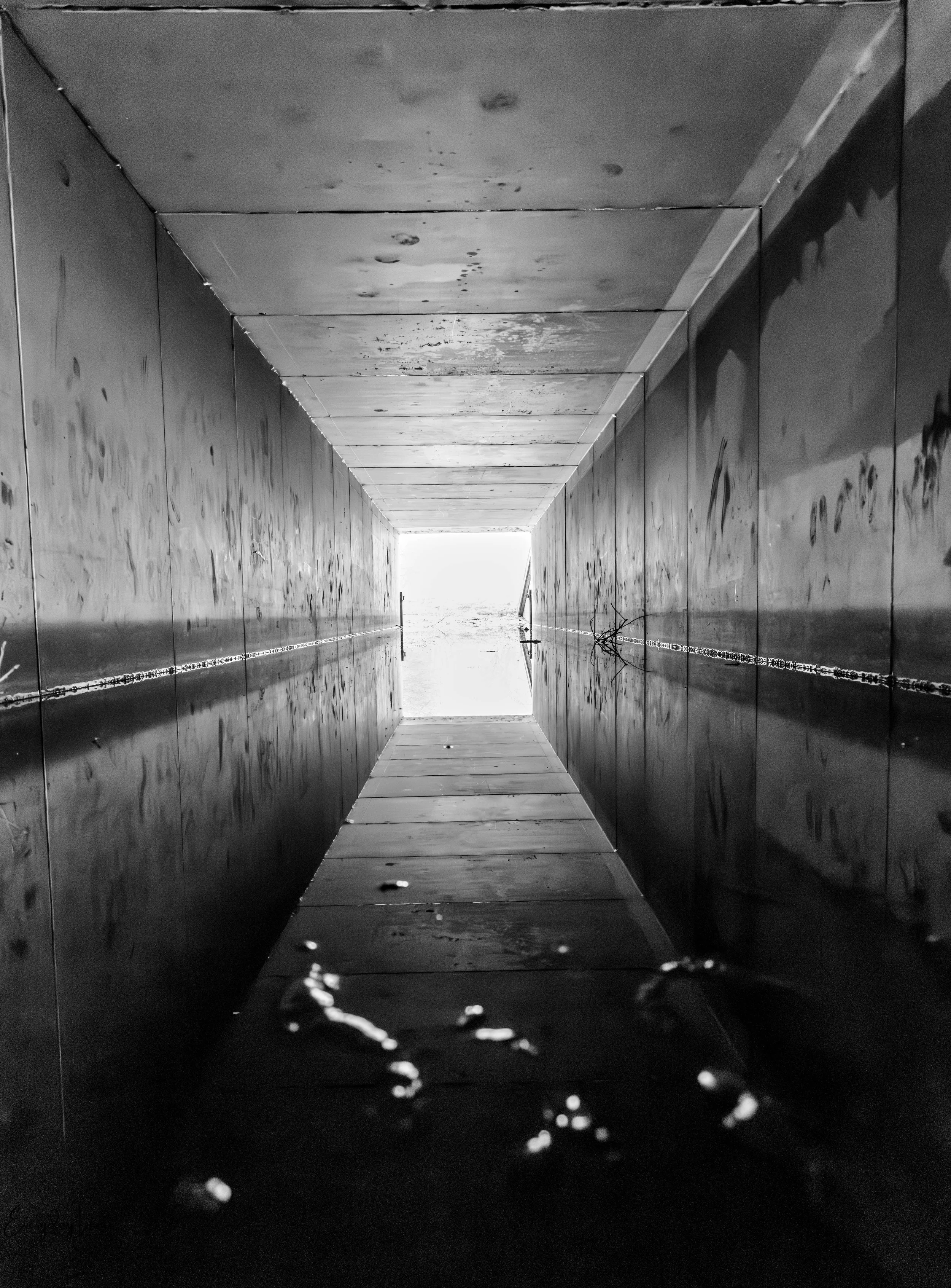 Looking down an enclosed narrow water passage with concrete sides and ceiling, with bright light at the far end and reflections of the passage on the water.