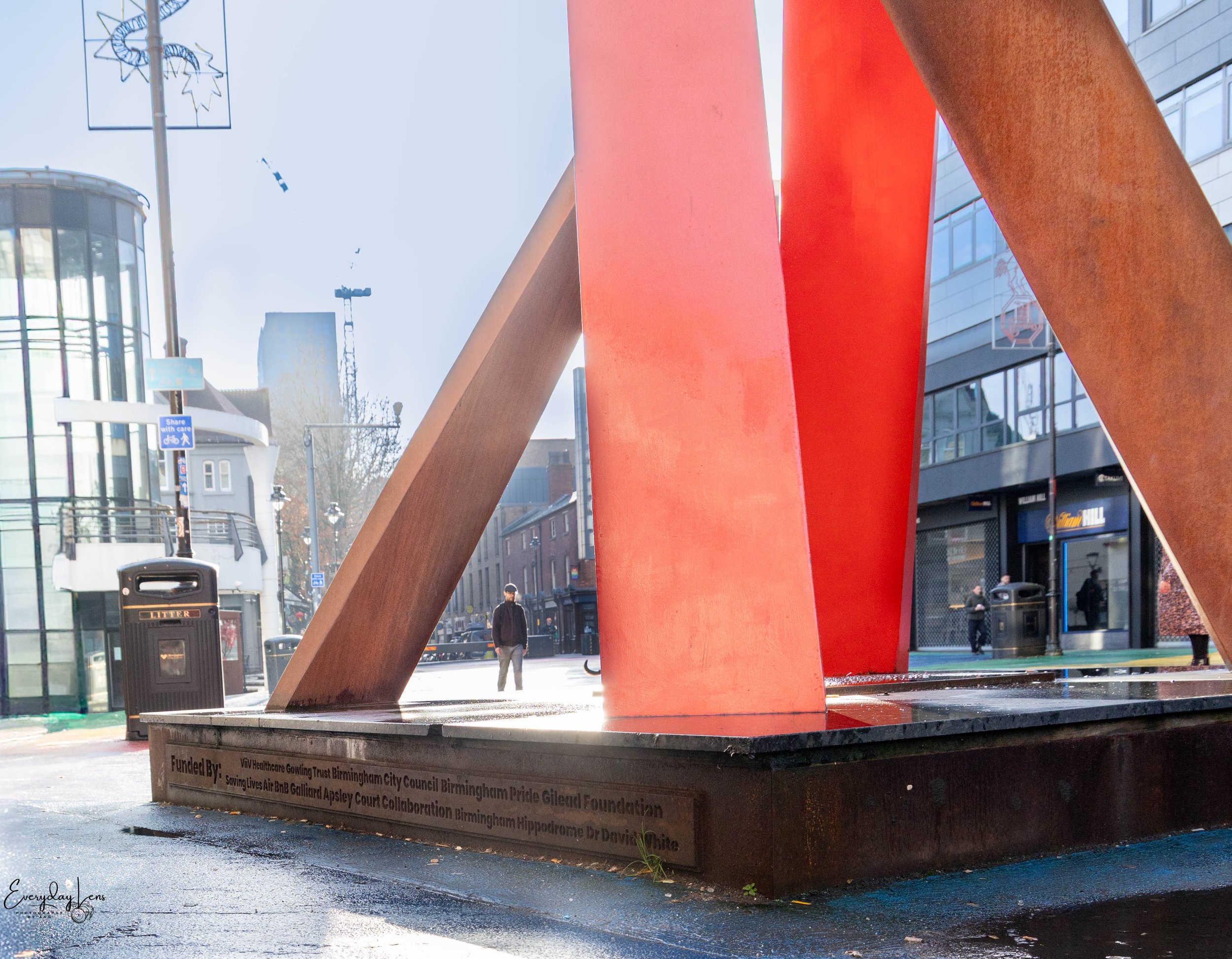 A large outdoor red sculpture with wooden and metallic elements on a city street, with pedestrians, buildings, and trash cans in the background.