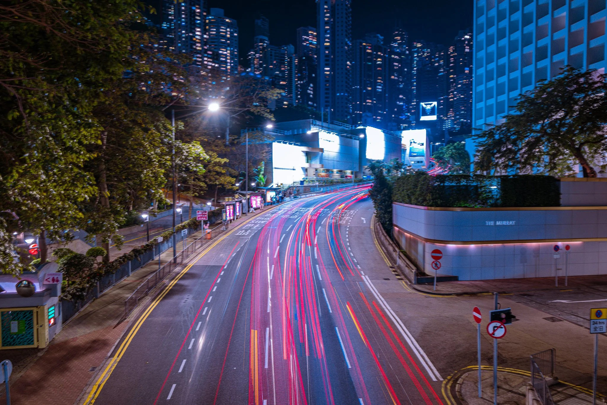 Nighttime city street with light trails from moving vehicles, skyscrapers in the background, and some greenery on the sides.