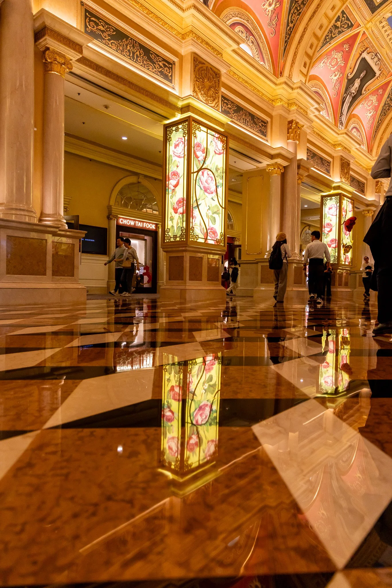 Interior of a luxurious hotel lobby with ornate ceilings and columns, illuminated decorative panels, and a polished marble floor reflecting the lighting and people walking through.