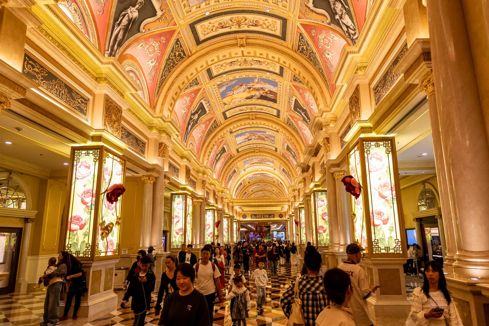 Interior view of a luxurious shopping mall corridor with ornate ceilings decorated with classical artwork, illuminated panels with floral designs, and a patterned marble floor, filled with people walking and shopping.