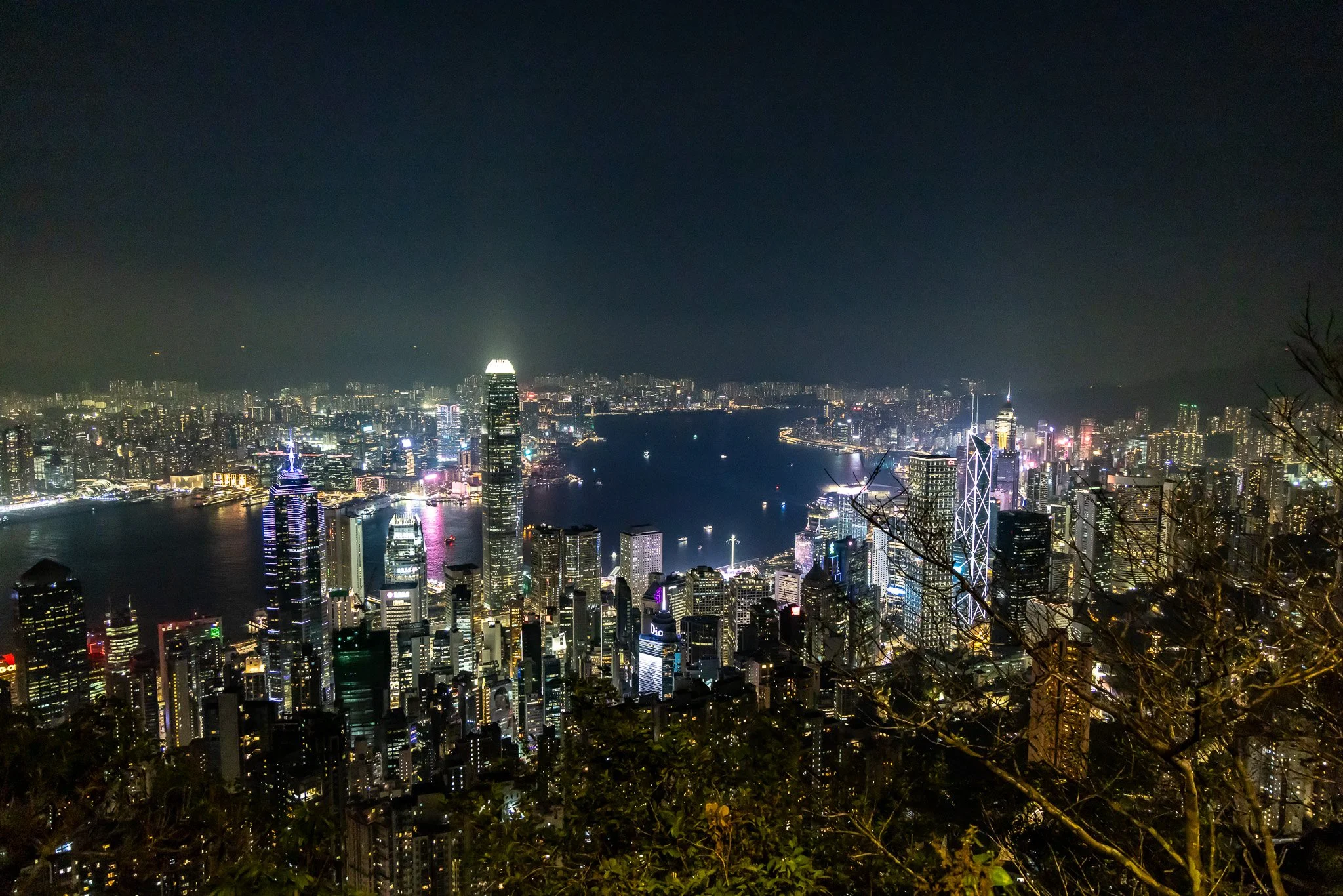 Nighttime cityscape of Hong Kong with illuminated skyscrapers, Victoria Harbour, and a dark sky.