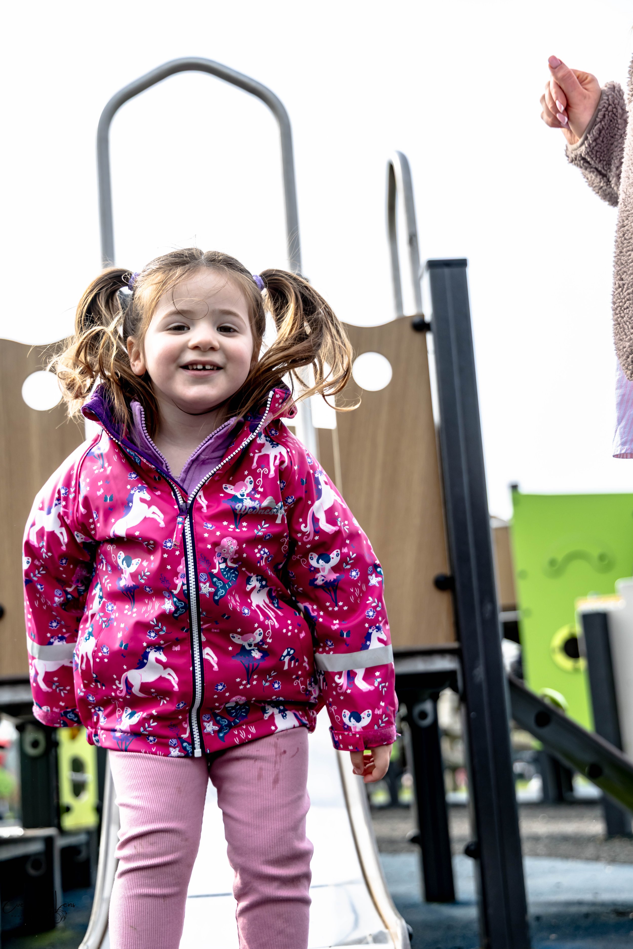 Young girl with brown hair in pigtails smiling at the camera, wearing a pink jacket with unicorn prints, standing near a playground slide.
