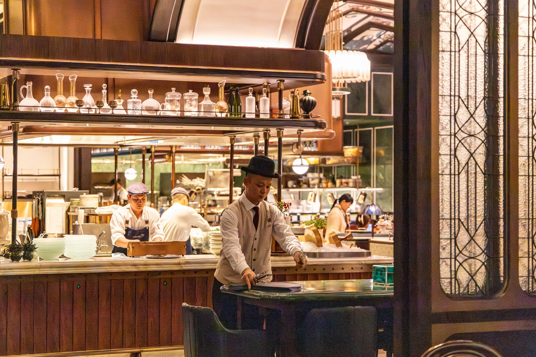 A young man in a white shirt, black tie, and black hat is cleaning a table in a restaurant or cafe, with a busy kitchen and other staff working in the background.