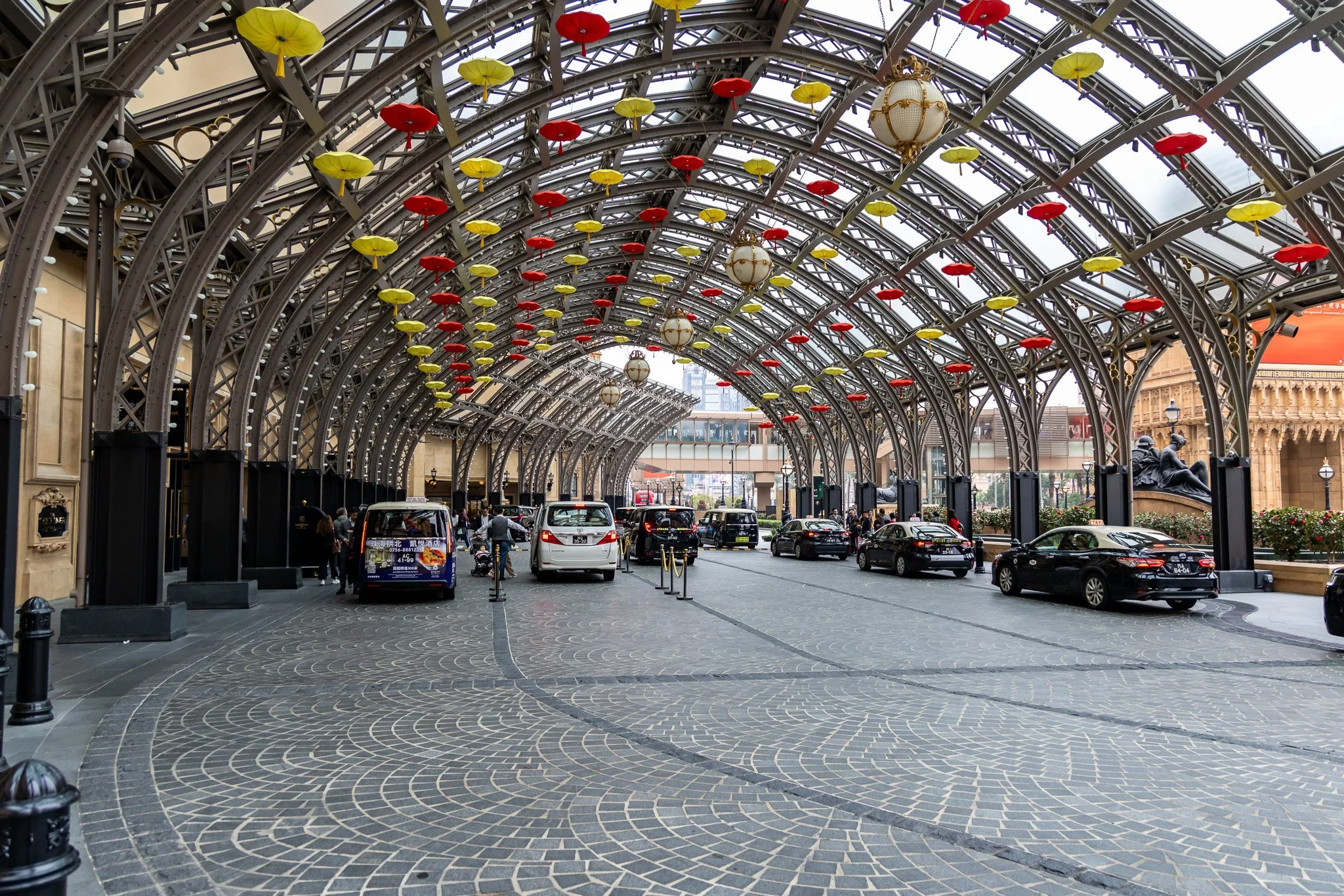 Covered drop-off area with iron arches decorated with red and yellow umbrellas hanging from the ceiling. Several cars and a few pedestrians are present.