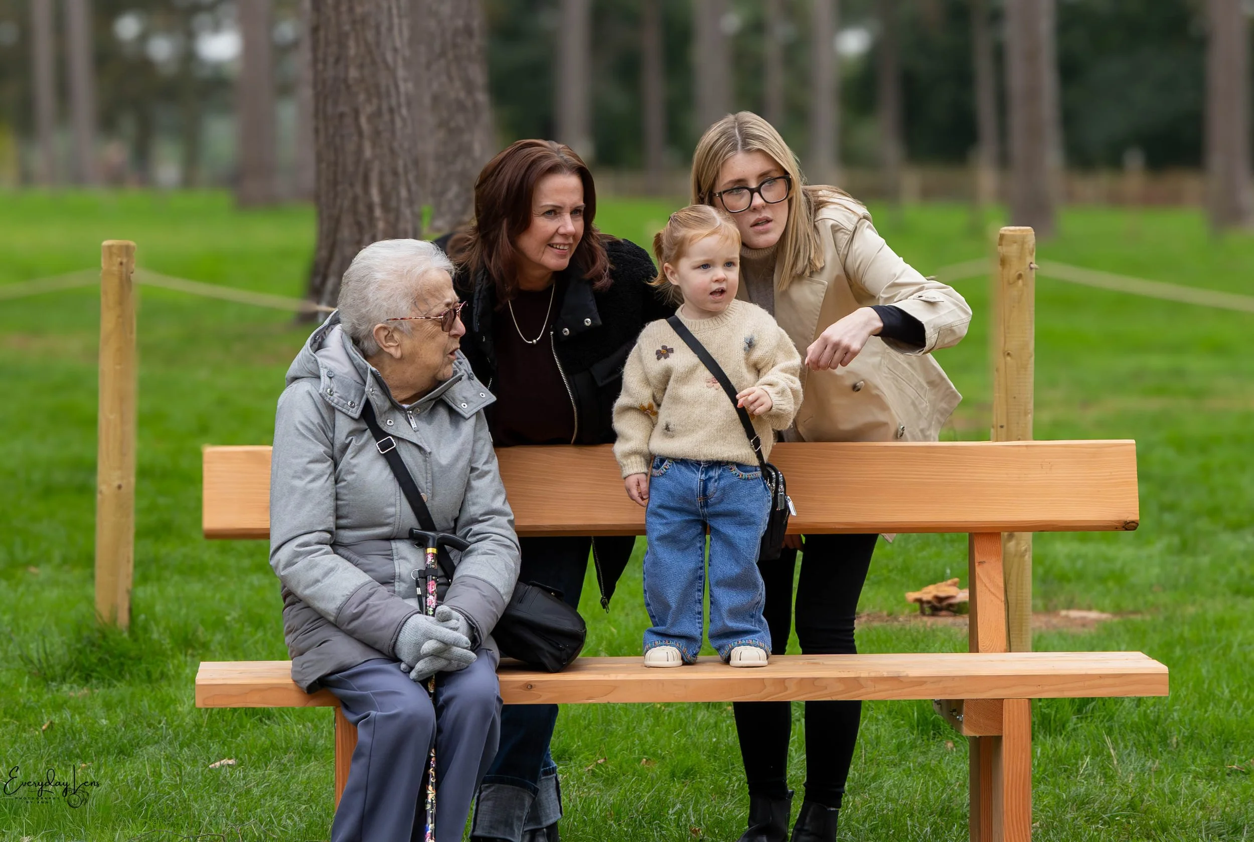Three women, including an elderly woman with white hair, three adult women, and a young girl, are on a wooden park bench outdoors surrounded by trees and green grass, with one woman pointing.