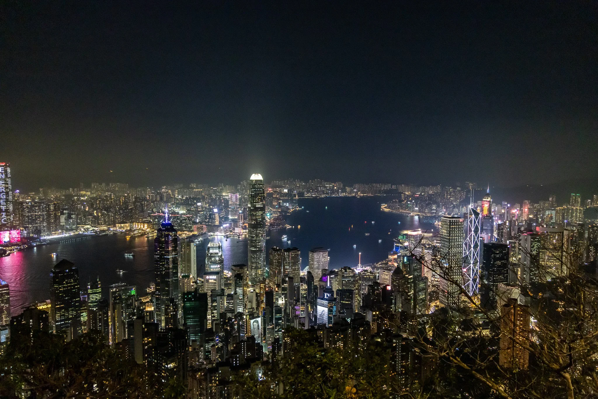 Nighttime aerial view of Hong Kong skyline featuring illuminated skyscrapers, Victoria Harbour, and surrounding mountains in the distance.